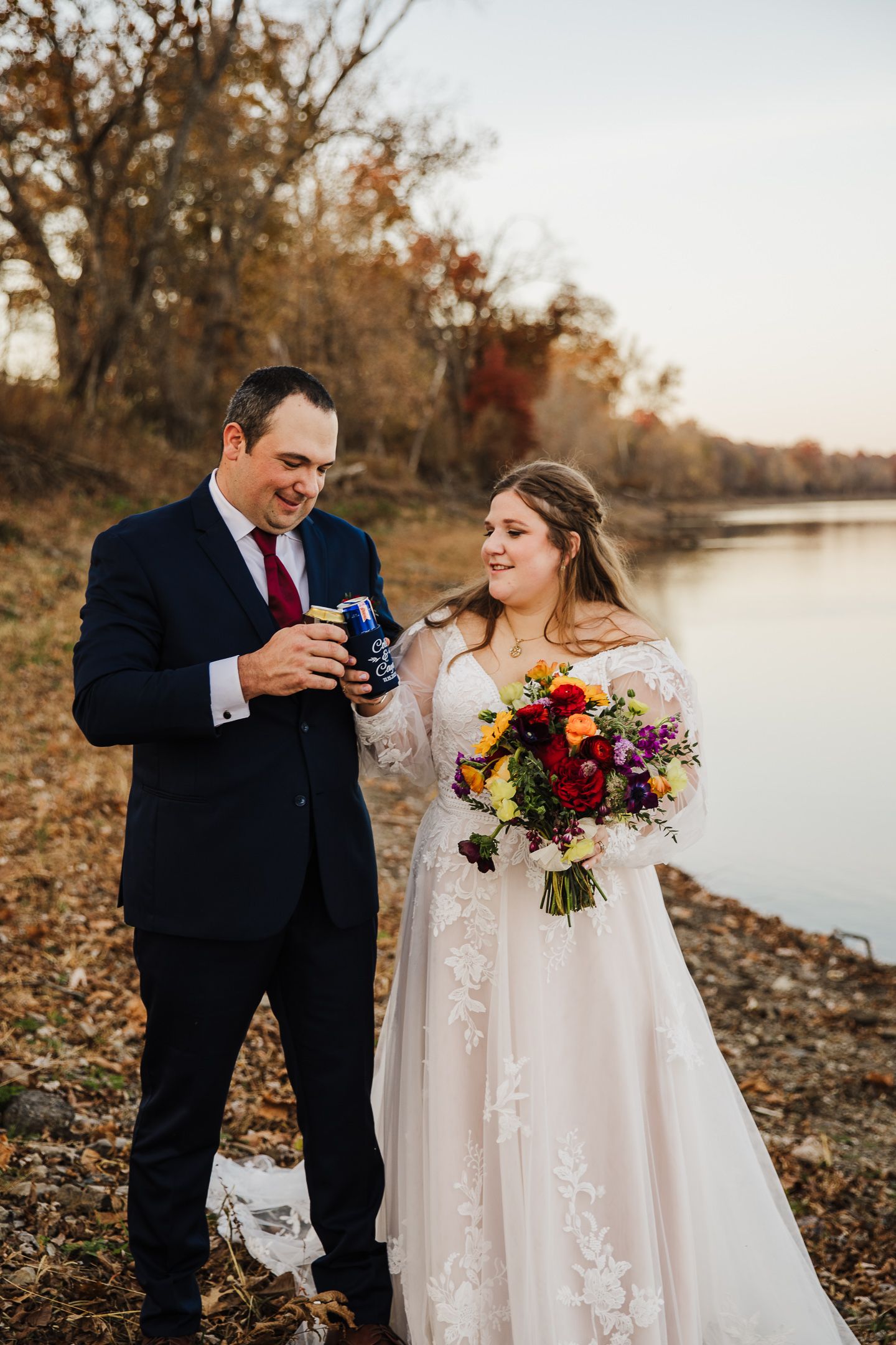 A couple shares a tender moment in autumn by a lake, with the bride holding a vibrant red and yellow bouquet.
