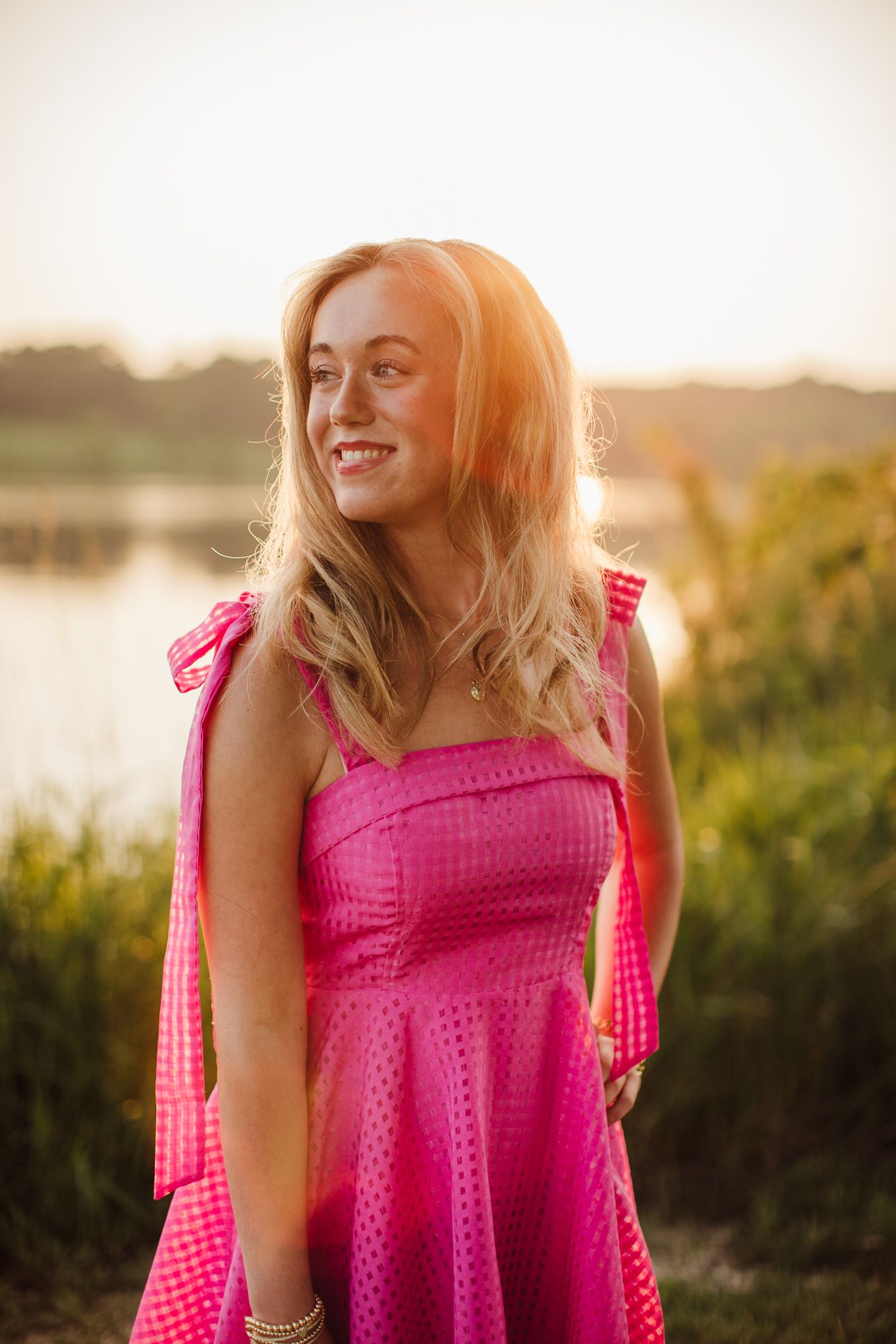 Person in bright pink dress laughing and posing in golden sunset light by water.