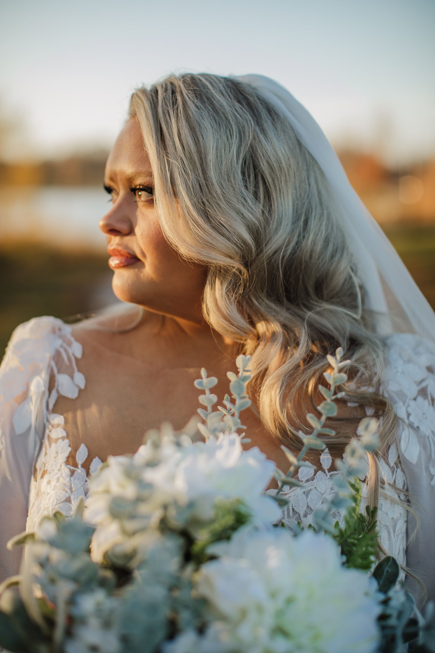A bride in a lace wedding dress poses at sunset with white flowers.