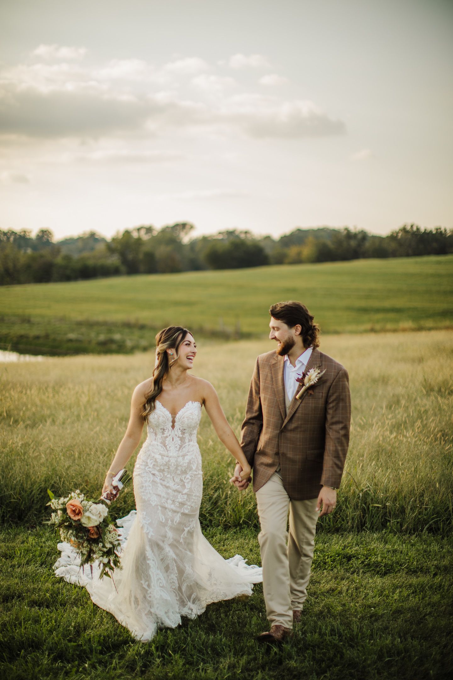 A couple walks through a golden meadow at sunset holding hands and carrying a floral bouquet.