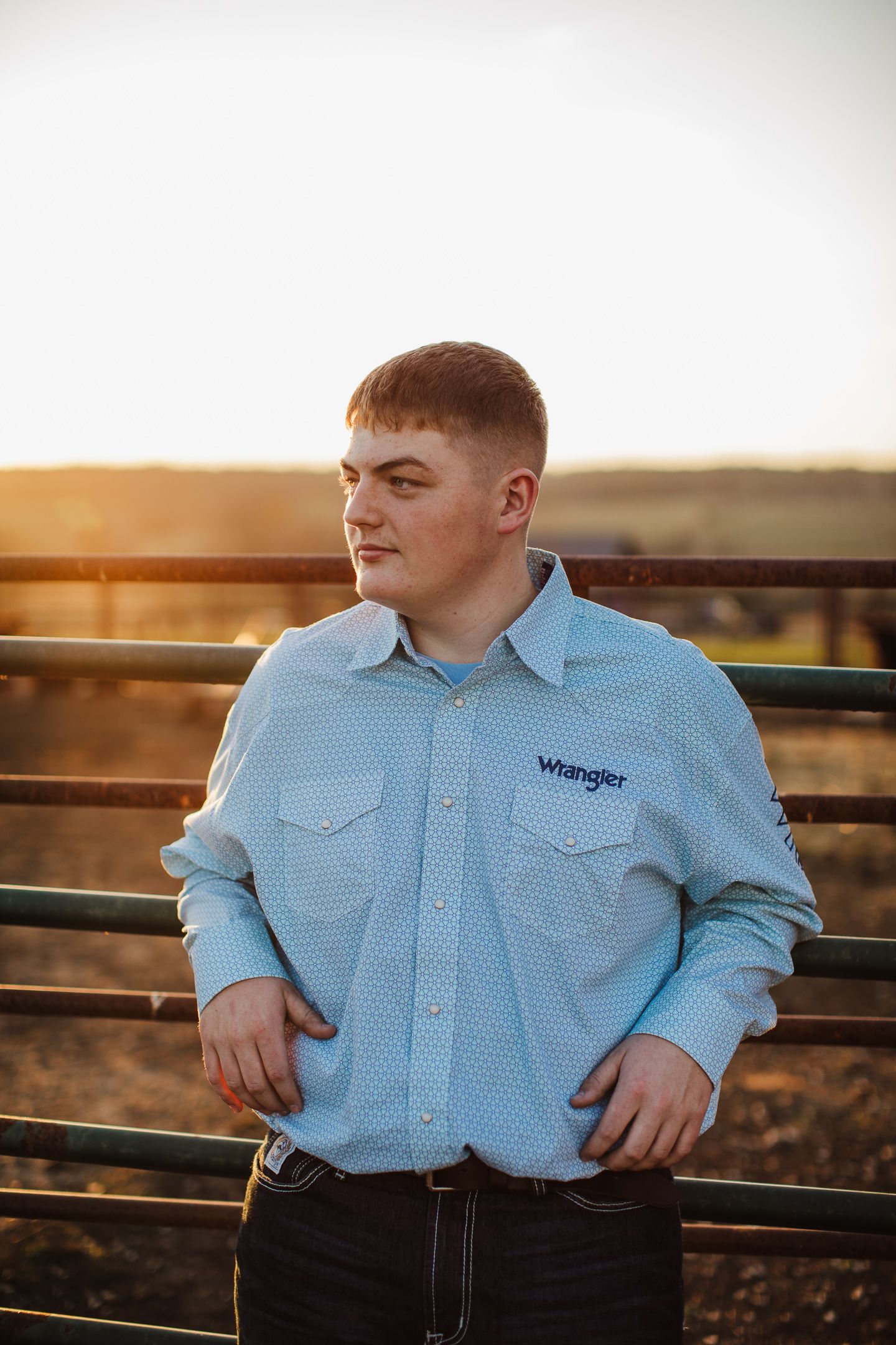 Person in light blue Wrangler western shirt poses against metal fence at sunset.