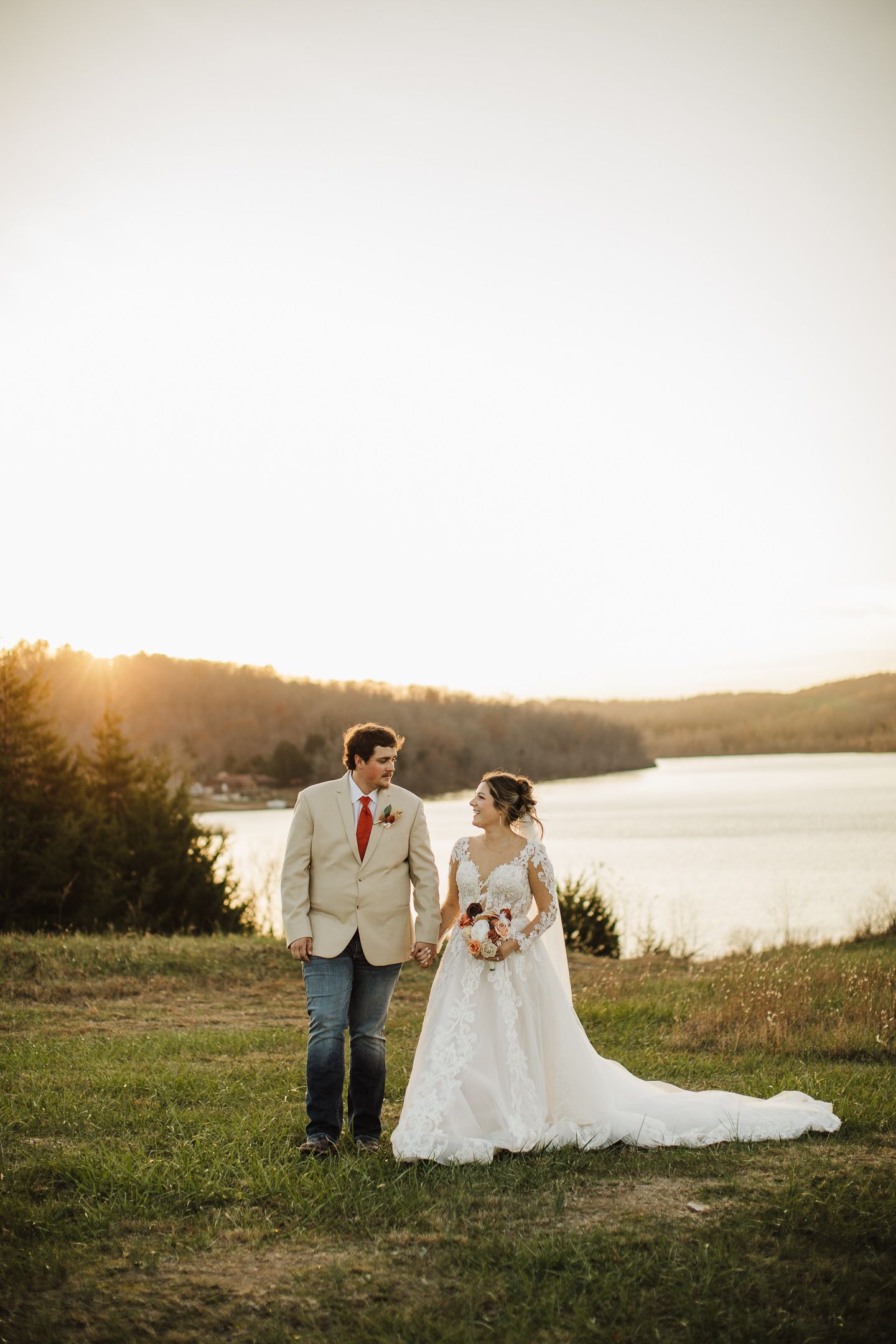 Wedding couple poses in scenic outdoor setting with lake and sunset in background.