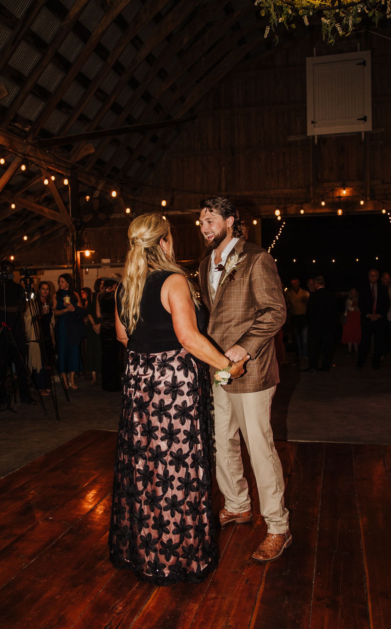 Couples dancing on wooden floor at dimly lit rustic wedding reception.