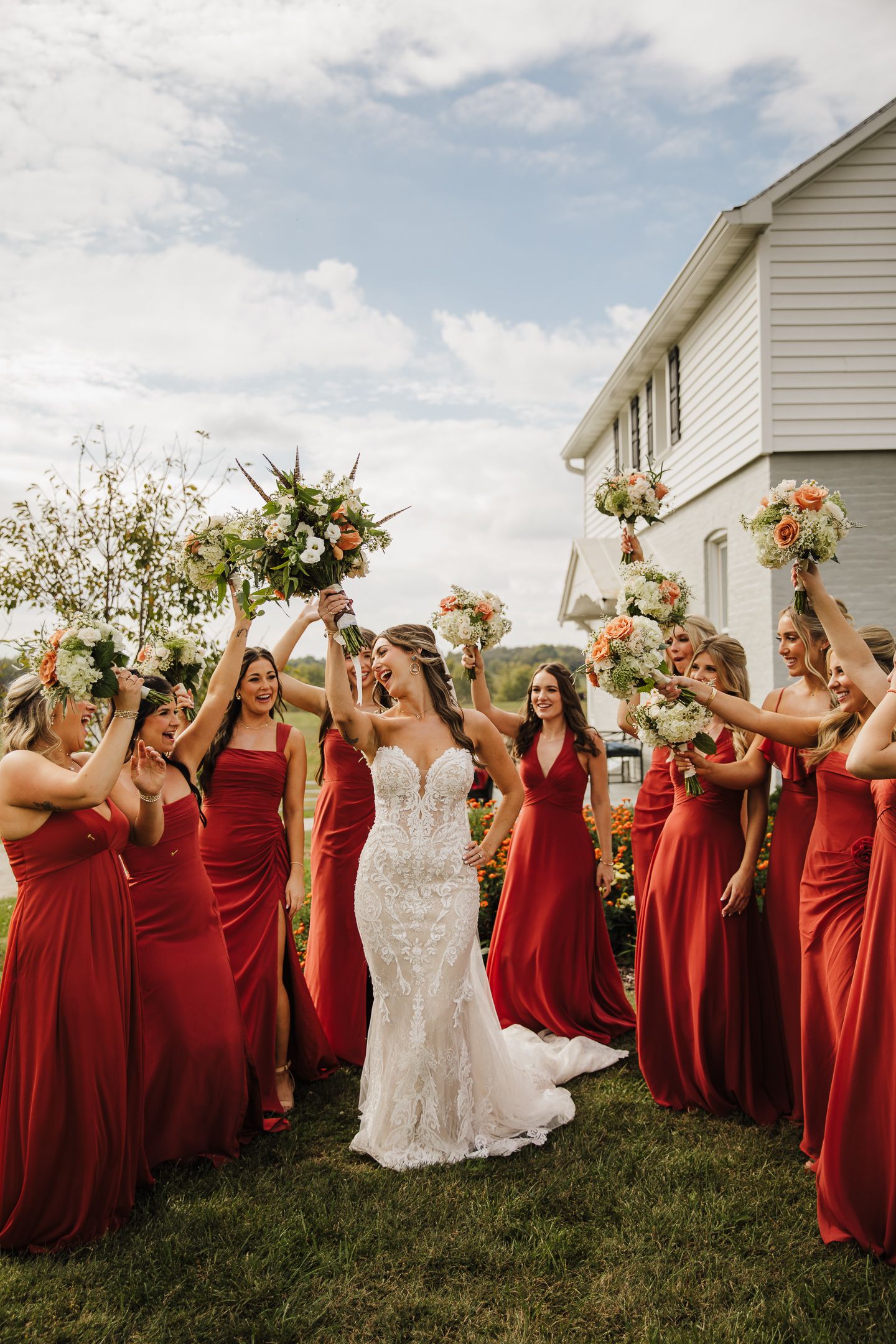Wedding party in red bridesmaid dresses posing outdoors with bride in white lace gown.