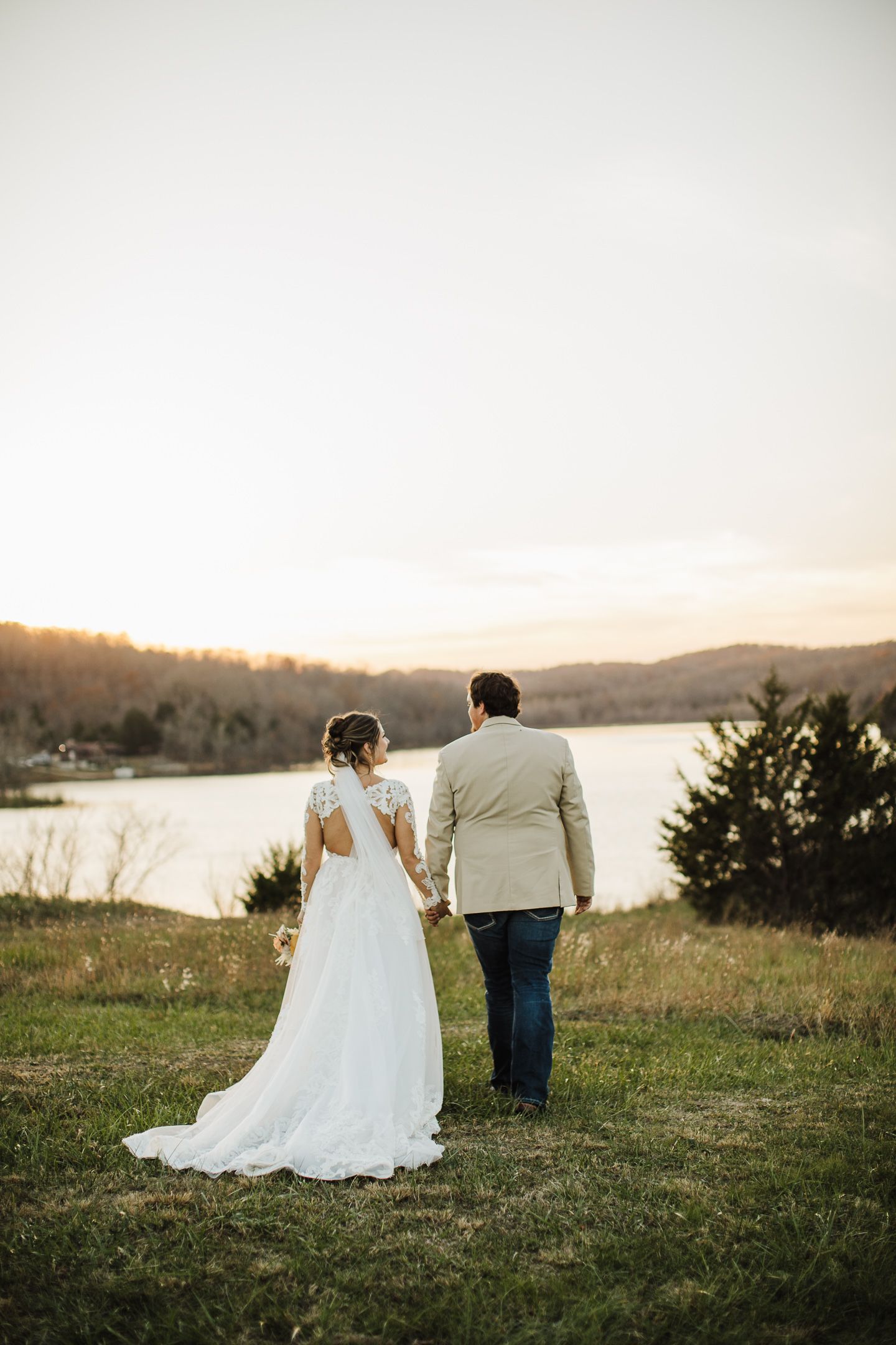 Newlyweds standing together overlooking a lake at sunset in romantic wedding attire.