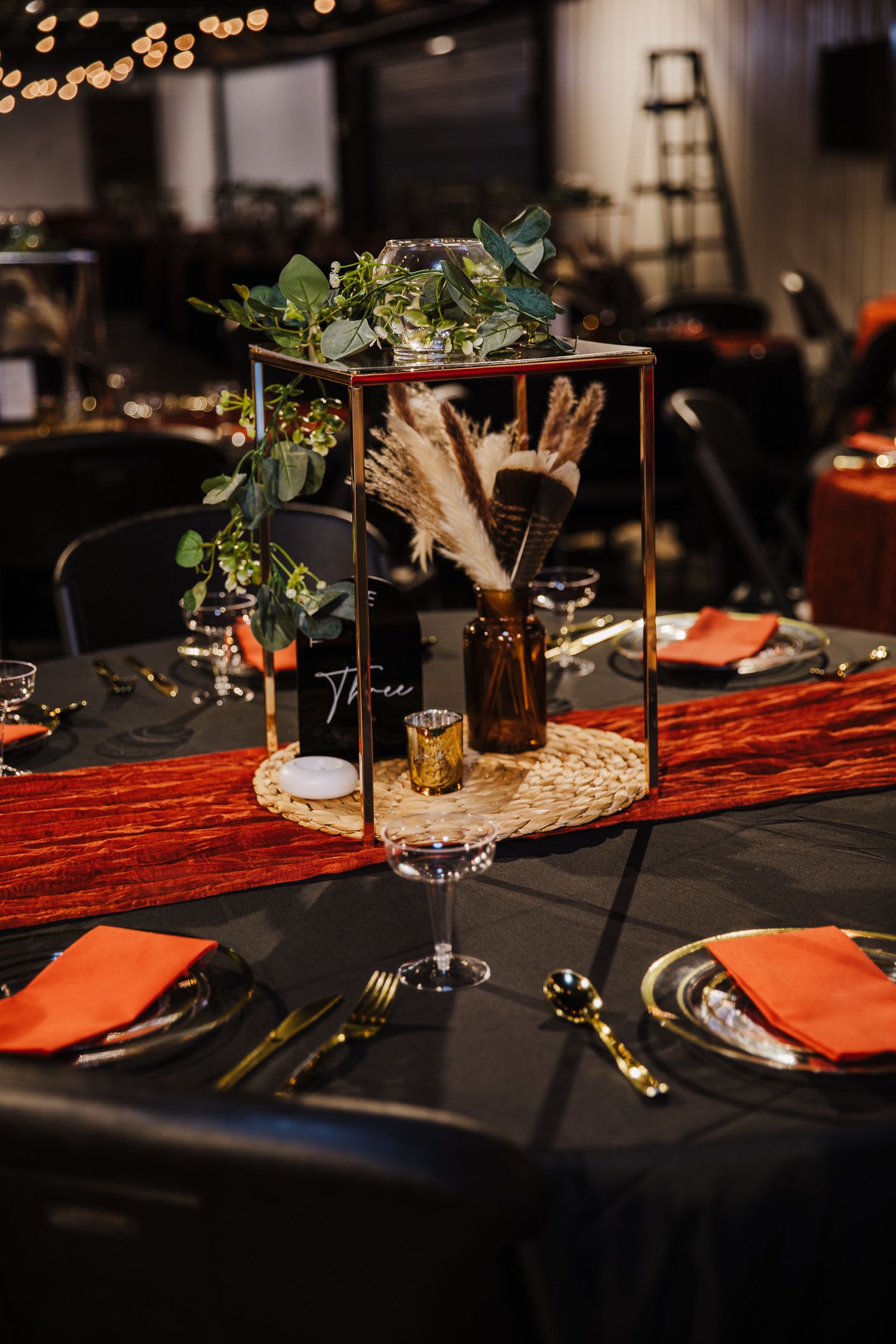 Elegant wedding table setting with black tablecloth, orange napkins, and greenery centerpiece in geometric frame.