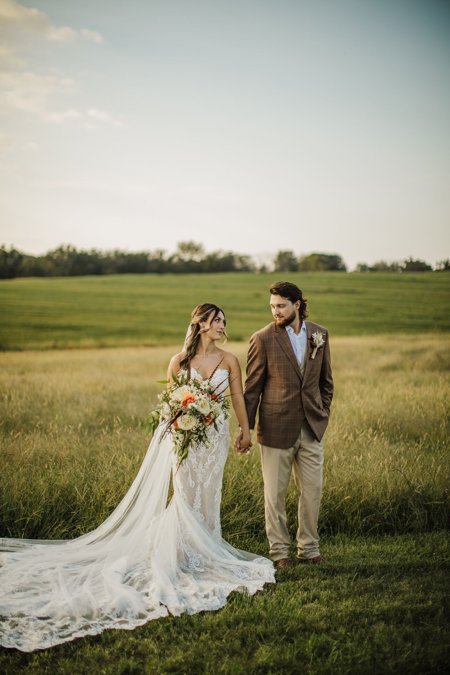 Bride in flowing lace gown and groom in brown suit pose in golden field at sunset.