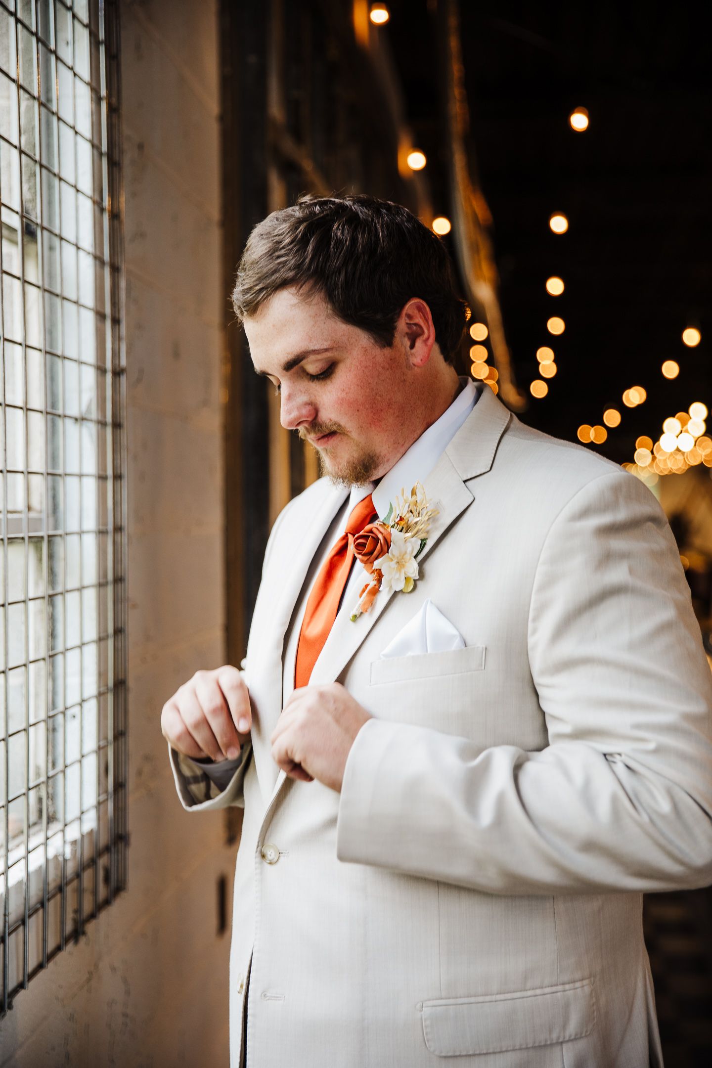 Groom in white suit adjusts tie against industrial window backdrop.