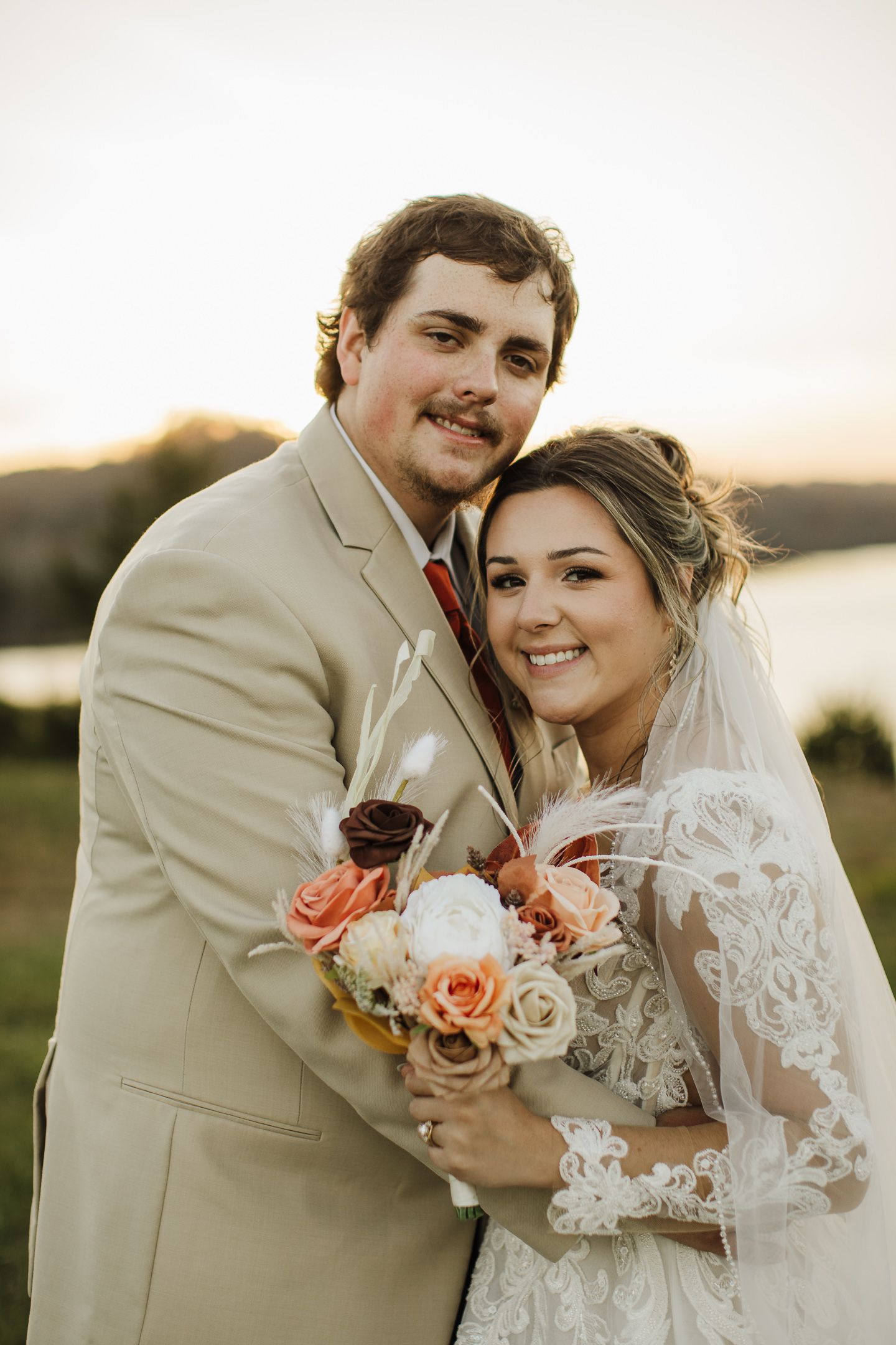 A romantic wedding photoshoot at sunset shows a couple embracing while holding a peach and coral rose bouquet.