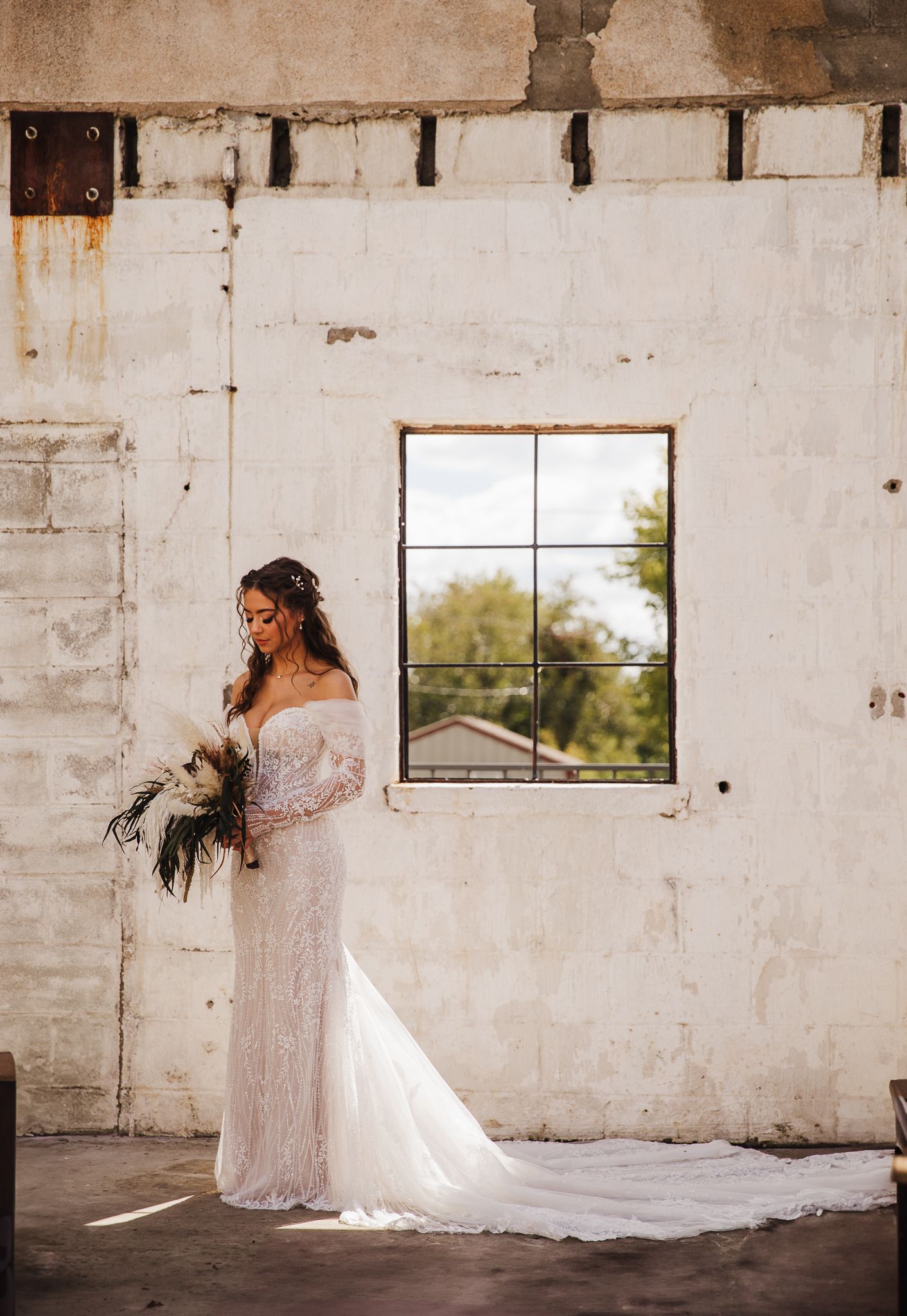 Bride in lace gown holding bouquet while standing in rustic room with window.