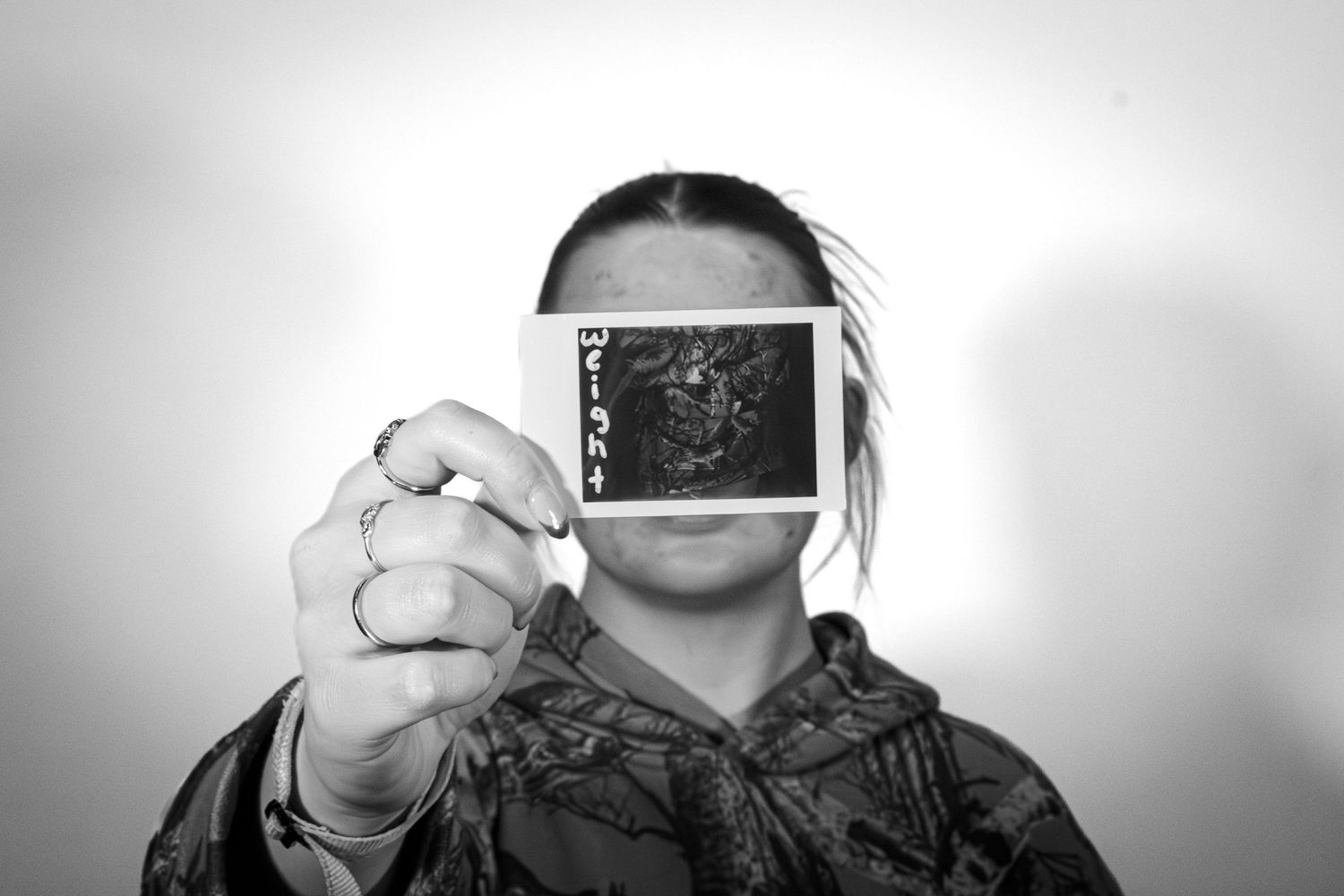 Person holding a Polaroid photo labeled Weight covering their face in a black and white portrait.