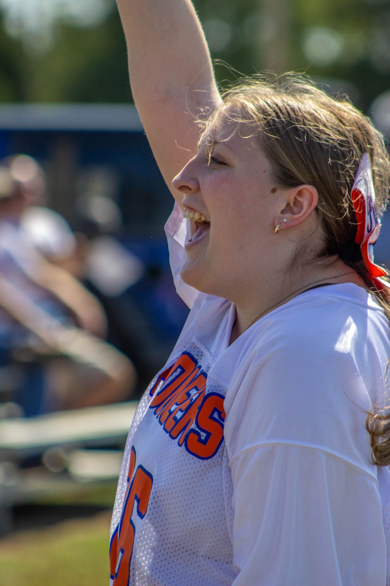 An enthusiastic young cheerleader in white and orange uniform raises her arm while cheering at a football game.