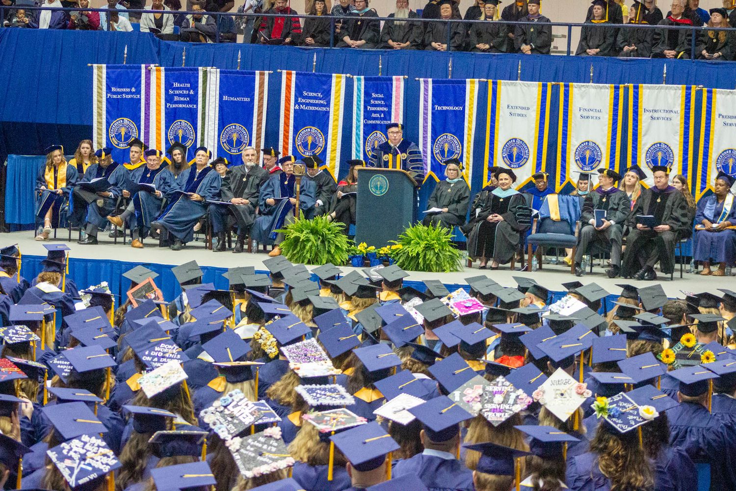 University graduation ceremony with rows of students in blue caps and gowns facing the stage.