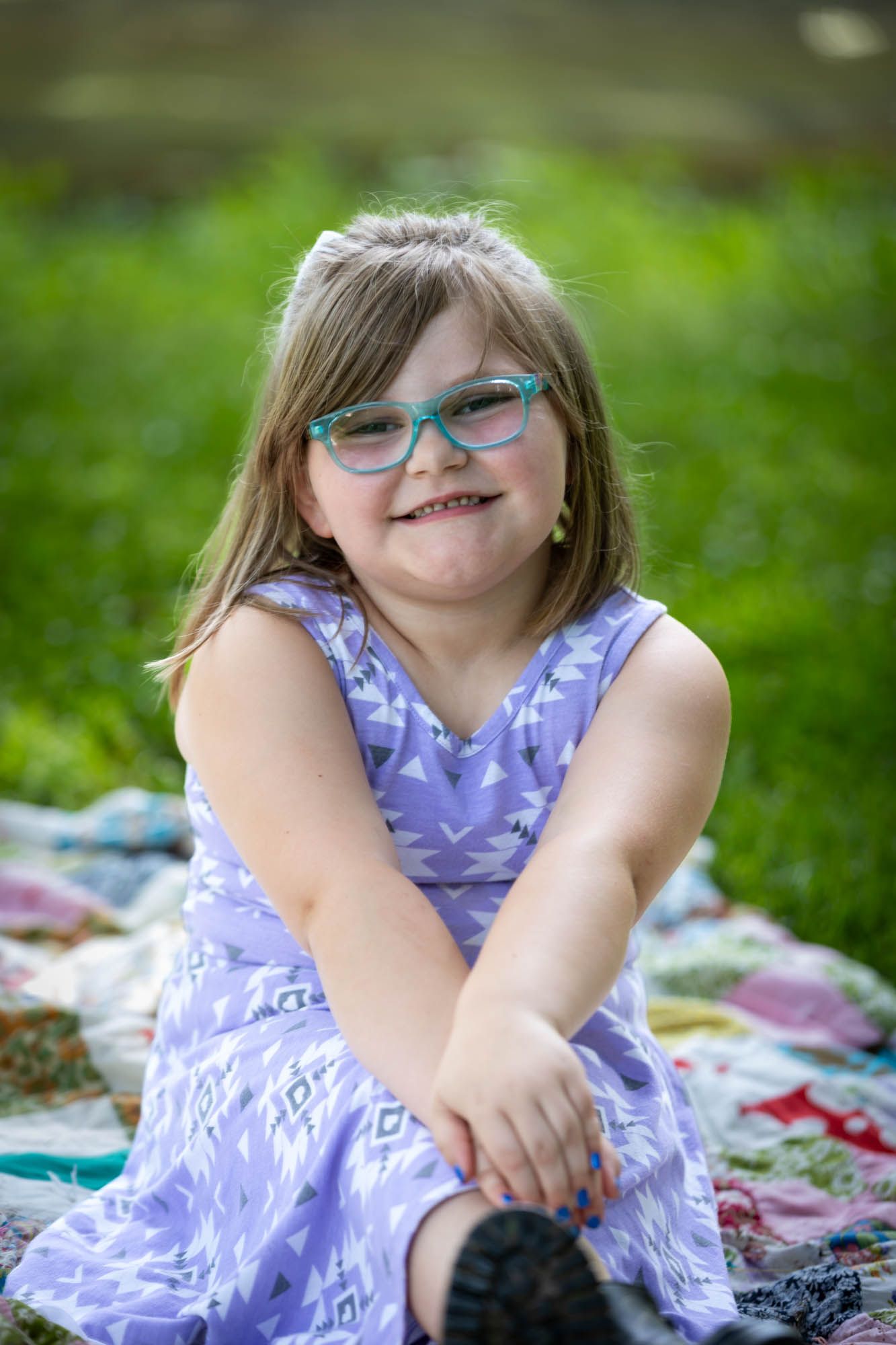 Young girl in purple dress and teal glasses smiling on colorful quilt outdoors.
