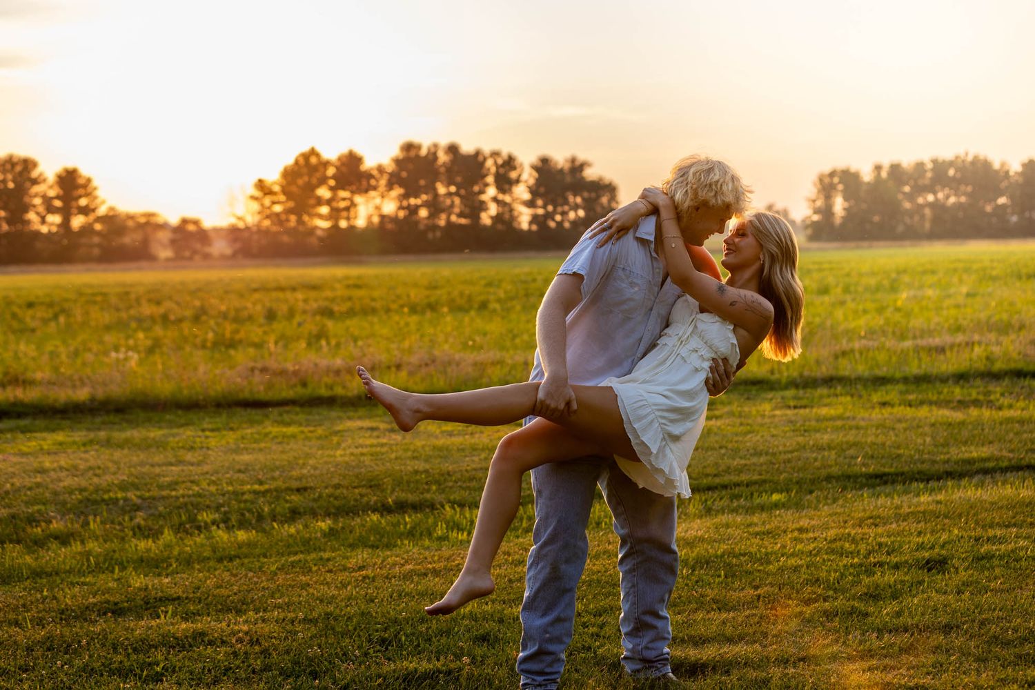 Couple sharing a romantic sunset moment in a green field, man lifting woman in a joyful embrace.