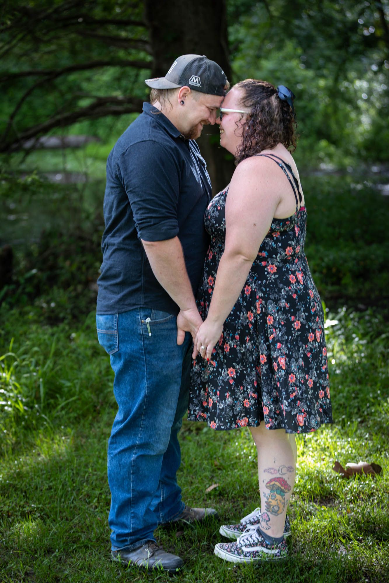A couple holds hands and touches foreheads romantically in a lush green park, woman in floral dress, man in cap and jeans.