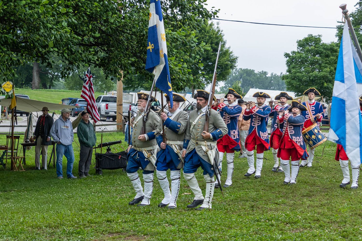 Colonial reenactors in period uniforms march with American and Scottish flags during a historical parade on a grassy field.