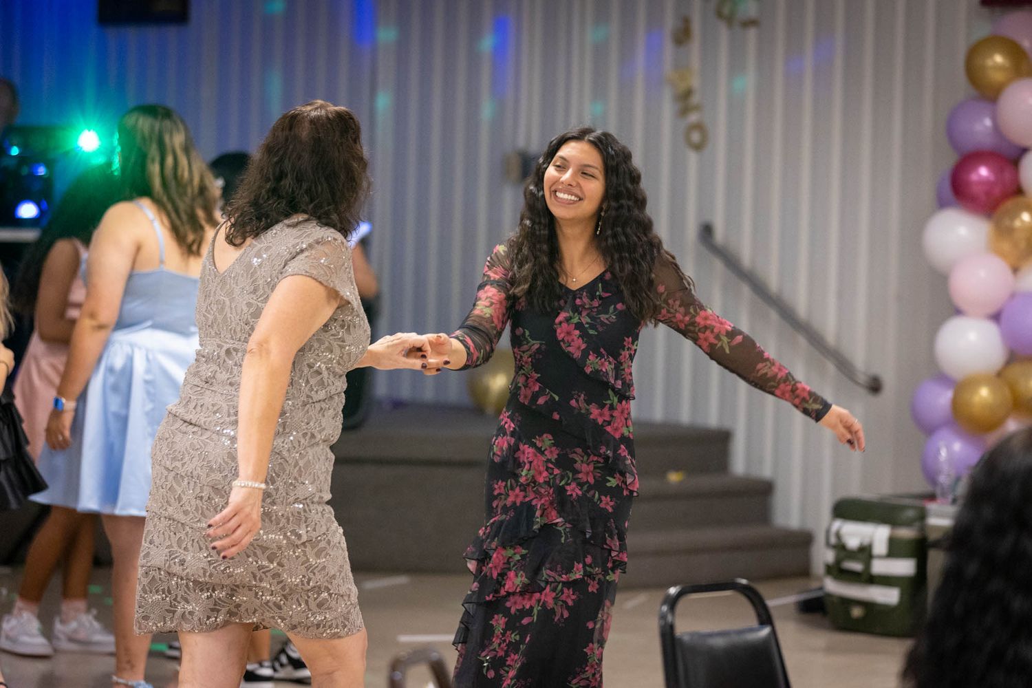 Two women dancing and smiling together at a quinceañera reception with balloons and colorful lighting.
