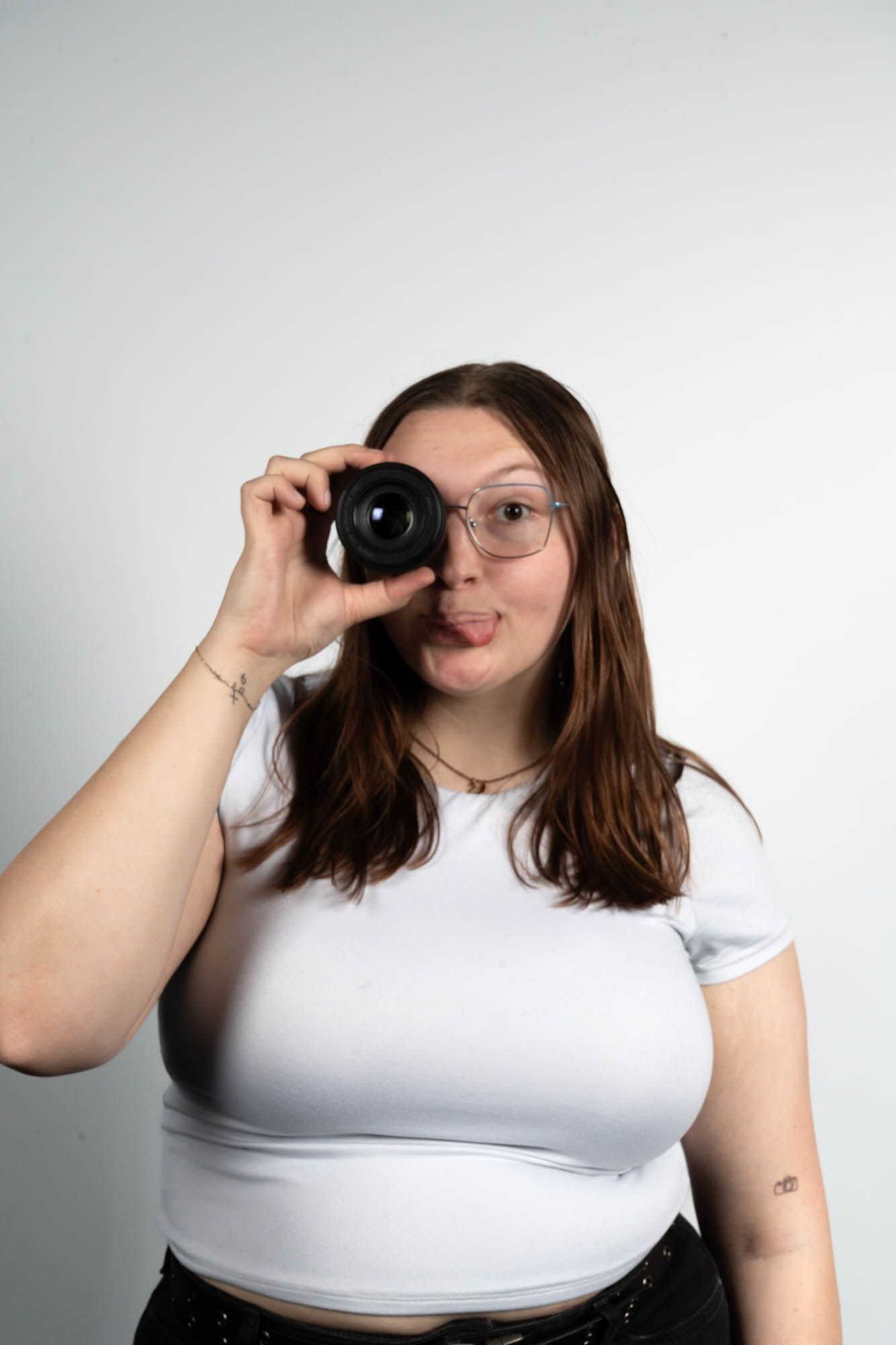 Young woman in white t-shirt making a funny face while holding a camera lens up to her eye.