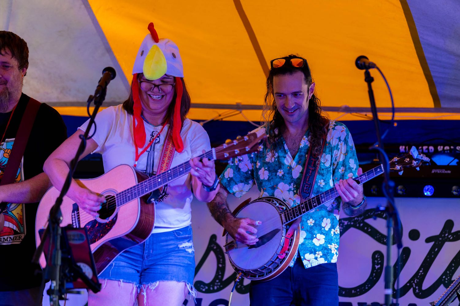 A band performs on stage at a lively outdoor music festival, one member wearing a chicken hat.
