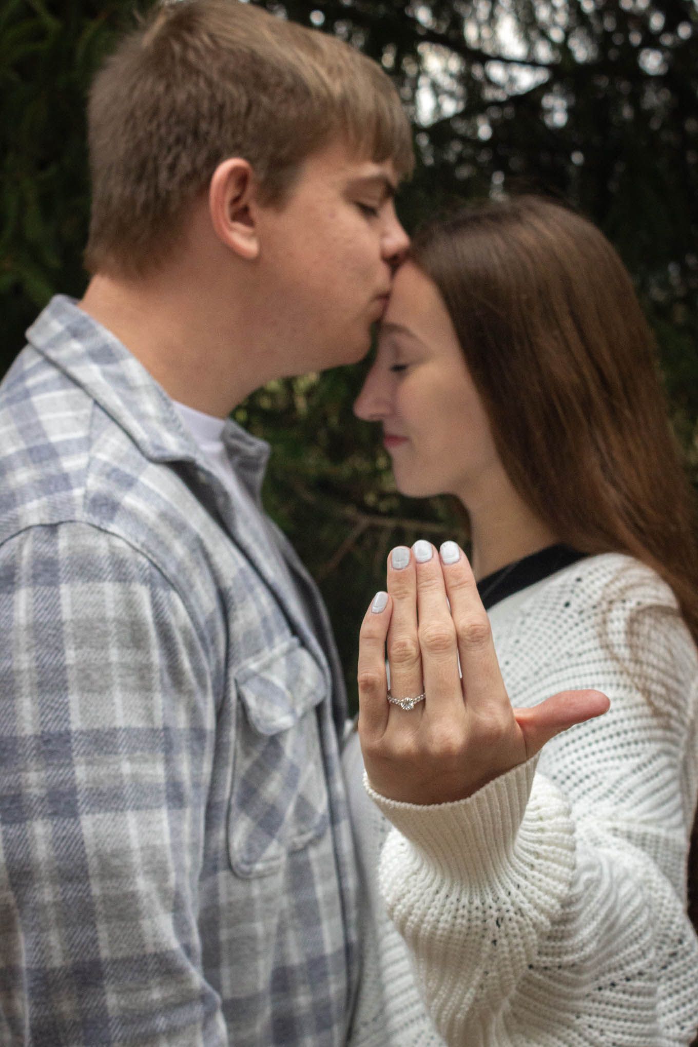 Engaged couple sharing forehead kiss while woman displays sparkling diamond engagement ring.