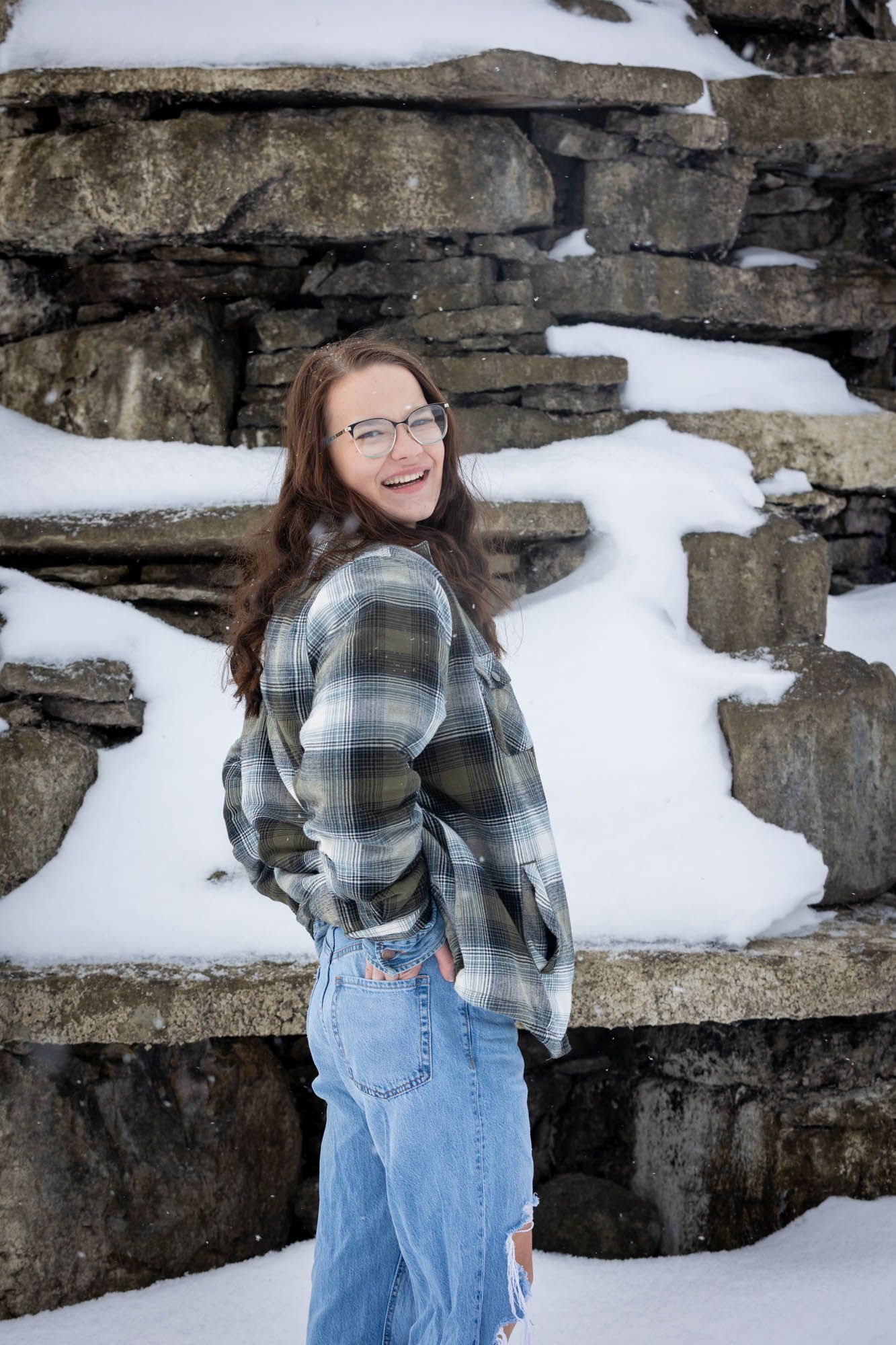 Young woman with glasses and long brown hair smiling in a plaid shirt against a snowy rock wall.