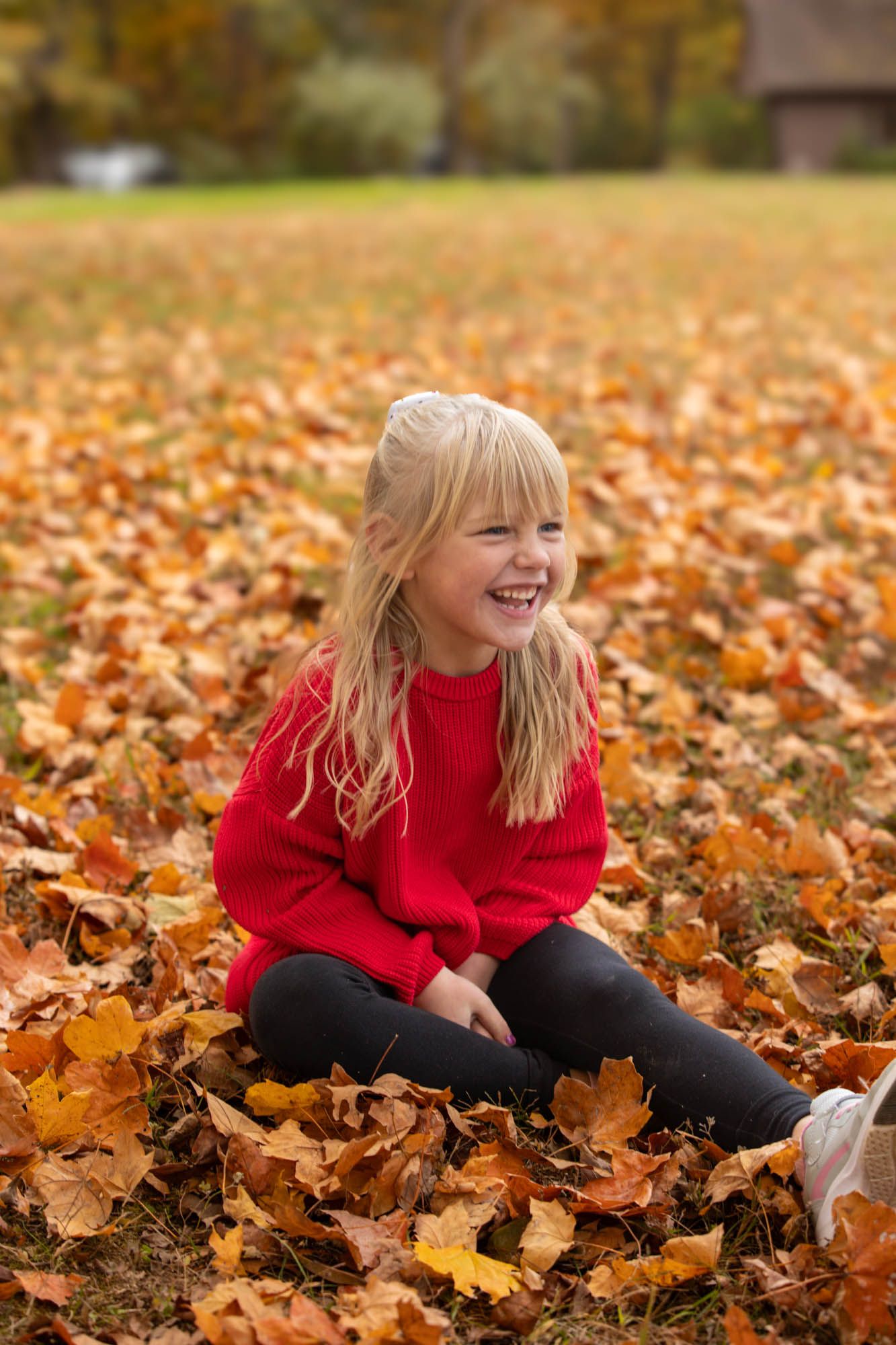 Joyful blonde girl in red sweater sits laughing among colorful fallen autumn leaves in a park.