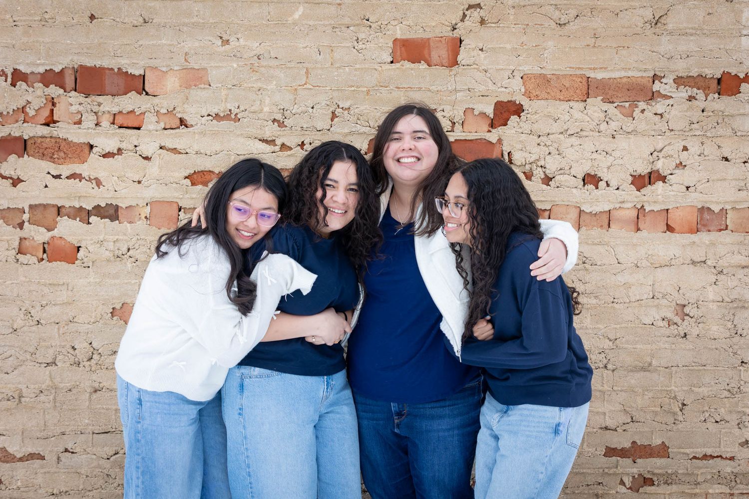 Four young women laughing and hugging each other in front of a rustic exposed brick wall.