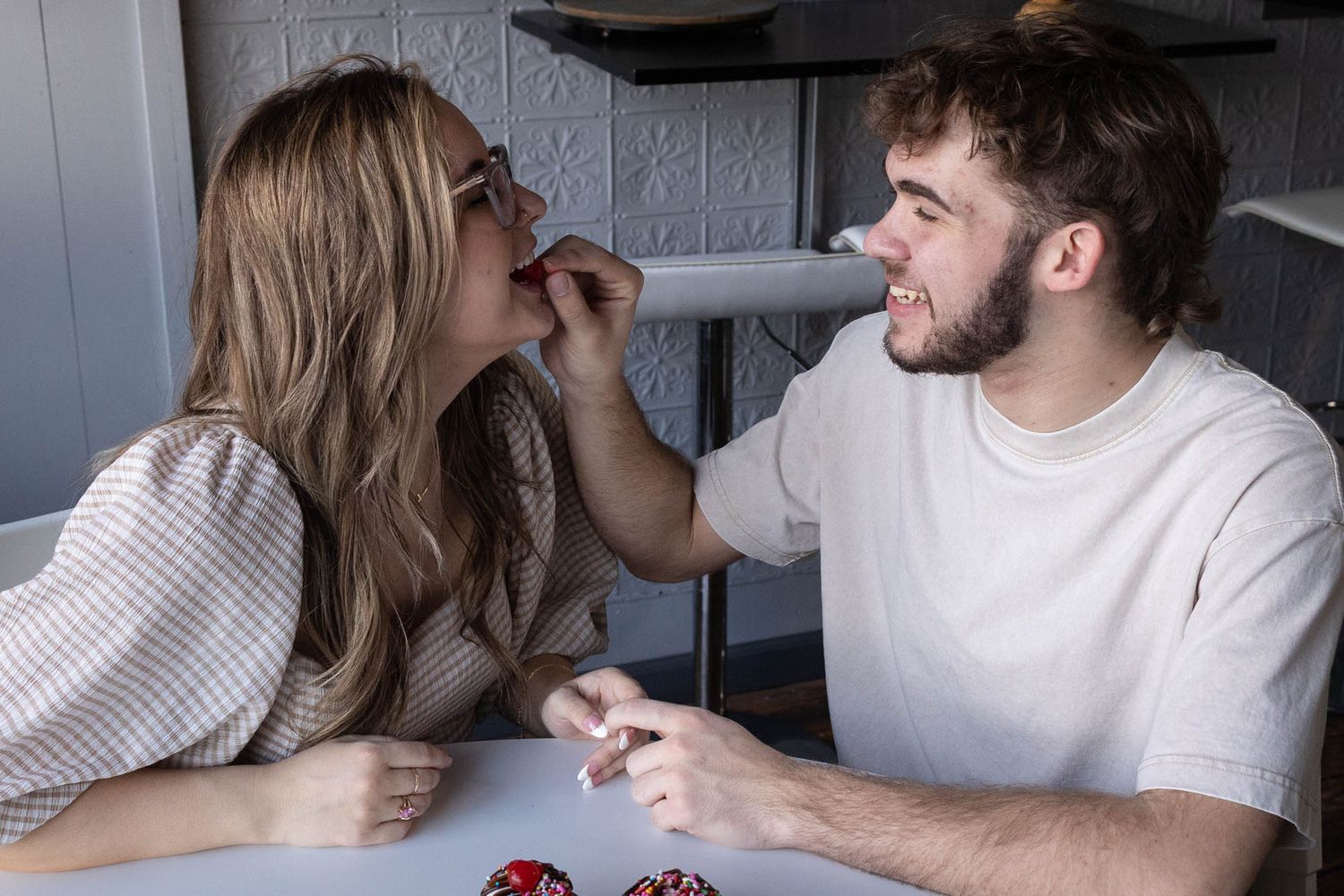 Playful couple feeding each other cherries at a white table, laughing and enjoying their date.