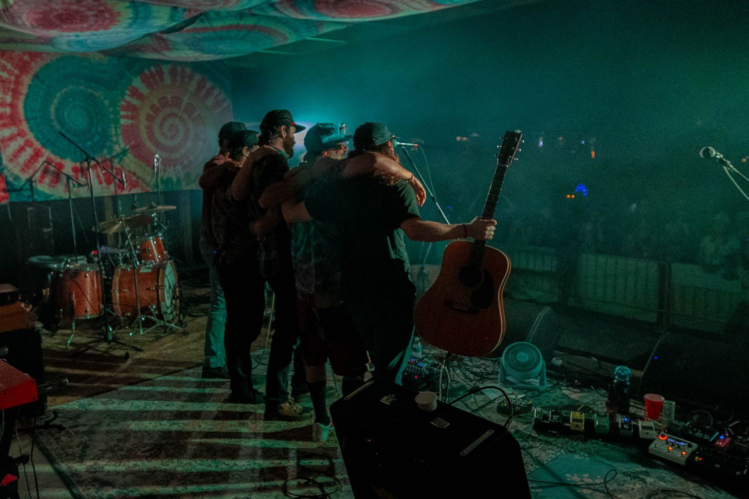 Band members share a group hug on stage beneath colorful psychedelic lighting during a concert.