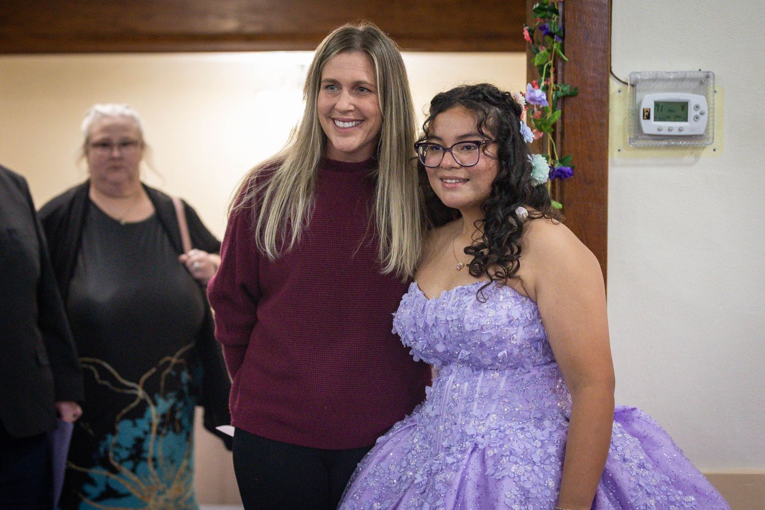 A young woman in a purple quinceañera gown poses with a smiling woman in a burgundy sweater at a celebration.