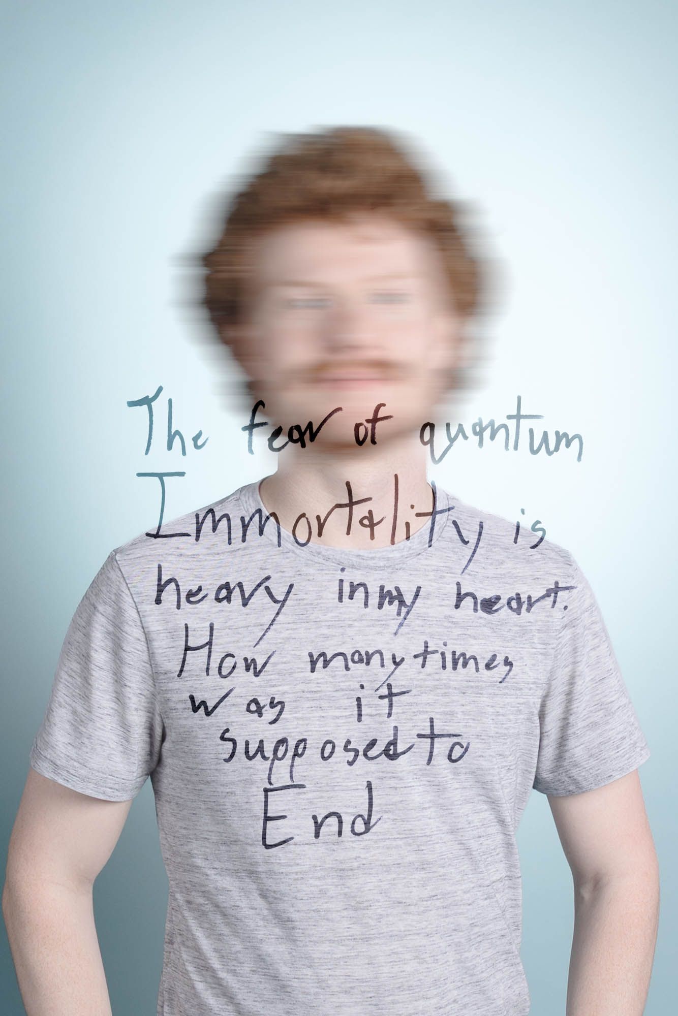 Blurred face of a redhead man in gray t-shirt with handwritten text about quantum immortality on light blue background.