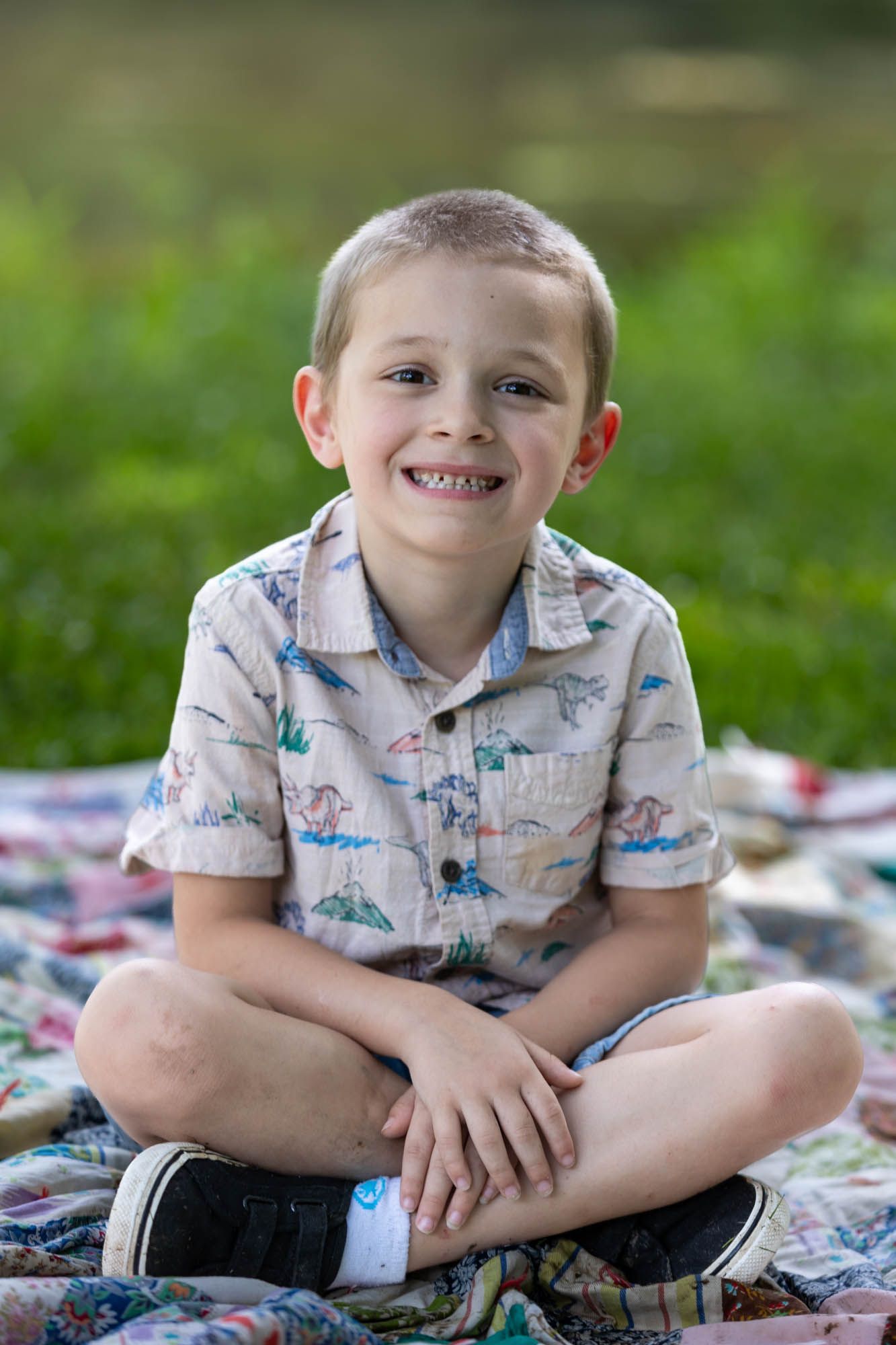 Smiling young boy sitting cross-legged on a colorful quilt outdoors, wearing a dinosaur print shirt.