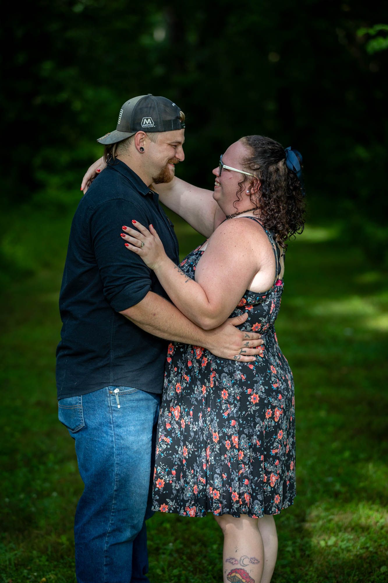 Couple embracing outdoors in a lush green forest, woman in floral dress and man in black shirt and cap.