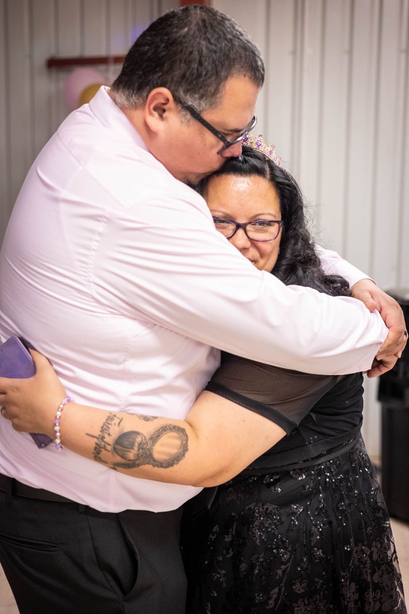 Man in white shirt shares emotional hug with woman in black dress at quinceañera reception.