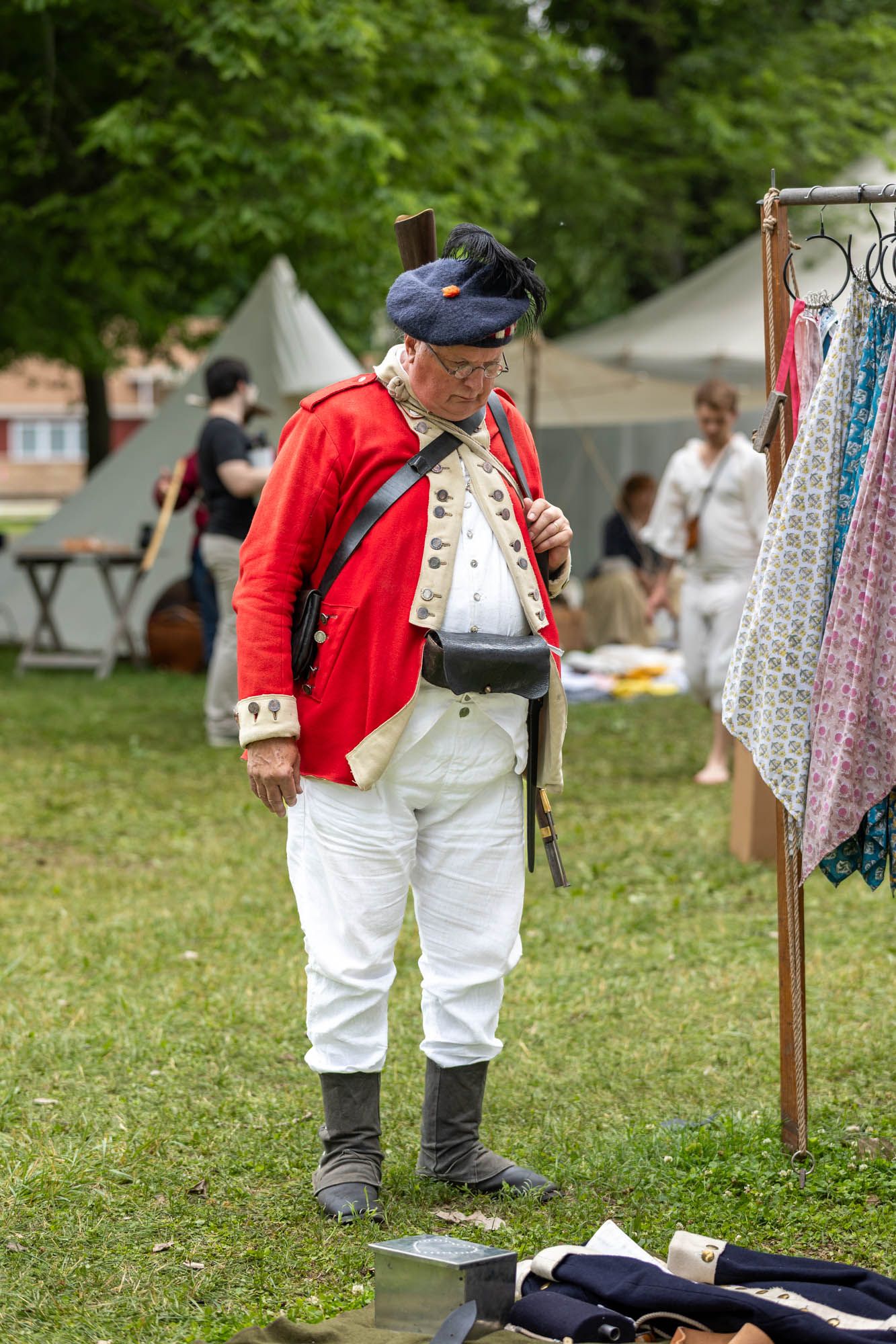 A male reenactor in a red British colonial military uniform examines items at an outdoor historical festival.