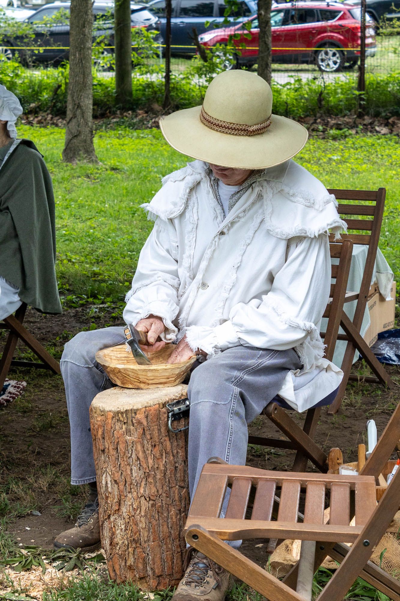 A historical reenactor in white colonial clothing uses a mortar and pestle on a tree stump at an outdoor event.