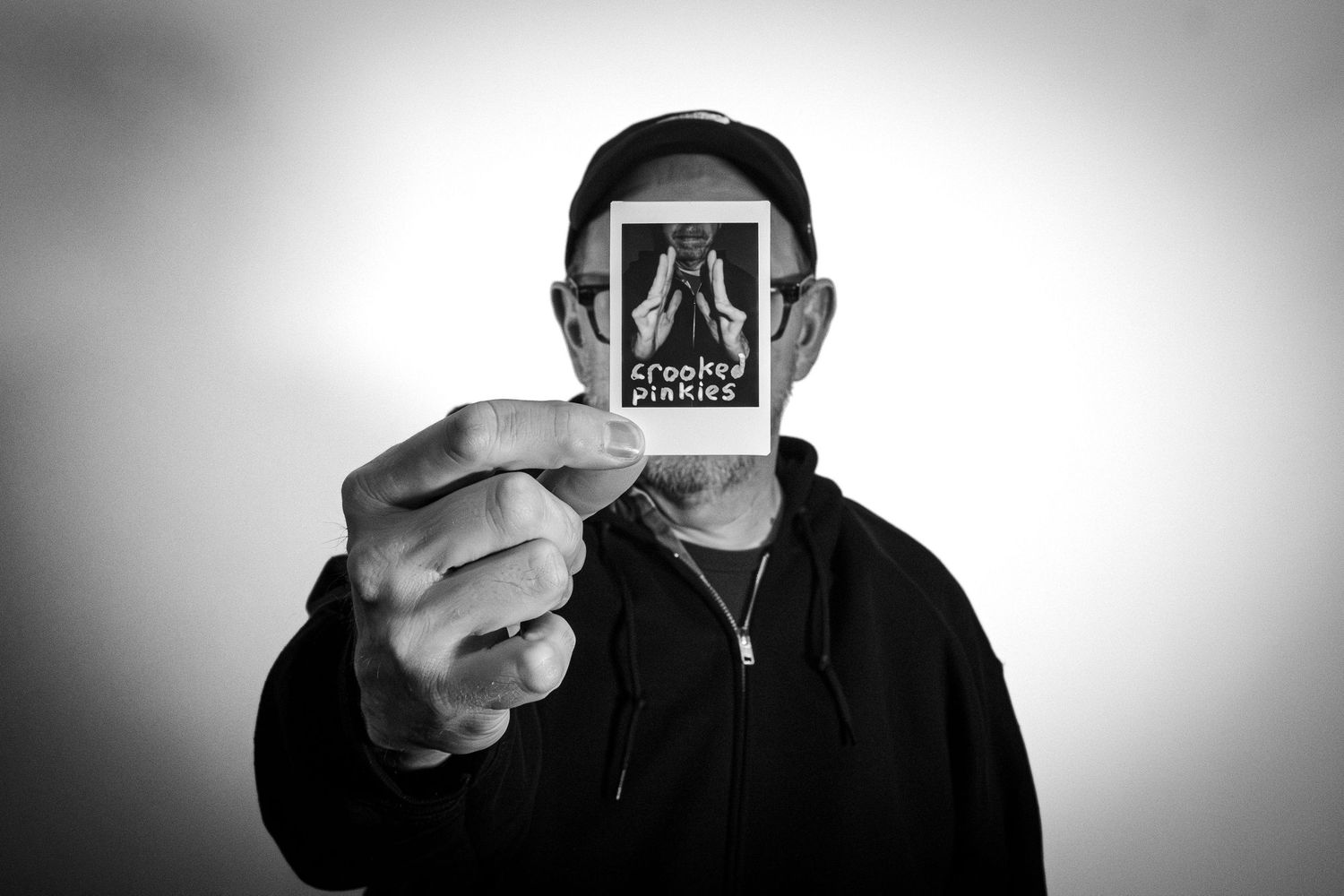 Man in a cap holding a Polaroid labeled Crooked Pinkies over his face in a black and white portrait.