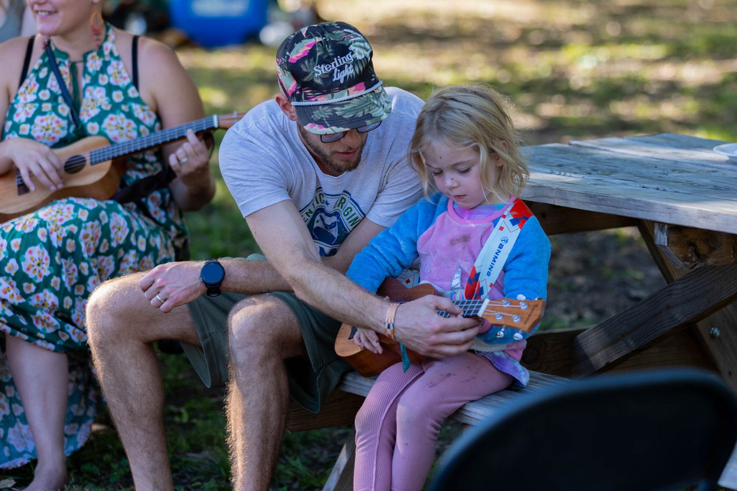 A man helps a young blonde girl play a colorful ukulele at an outdoor picnic table.