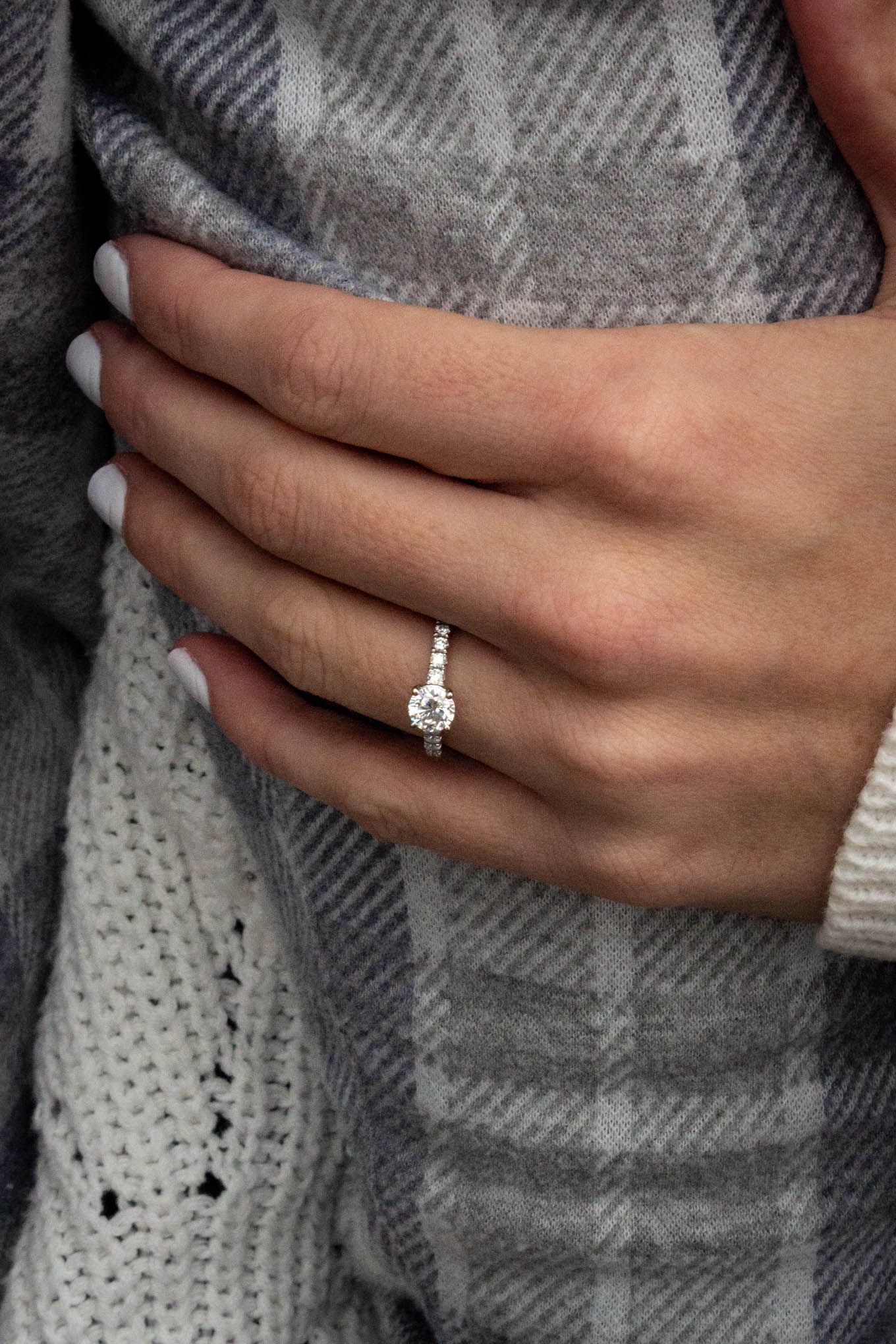 Close-up of a diamond halo engagement ring on a woman's hand against a plaid fabric background.