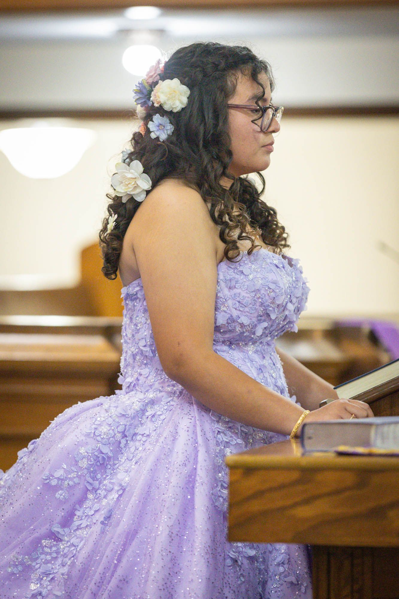 Teen girl in purple lace quinceañera dress with floral hair accessories stands at church podium.