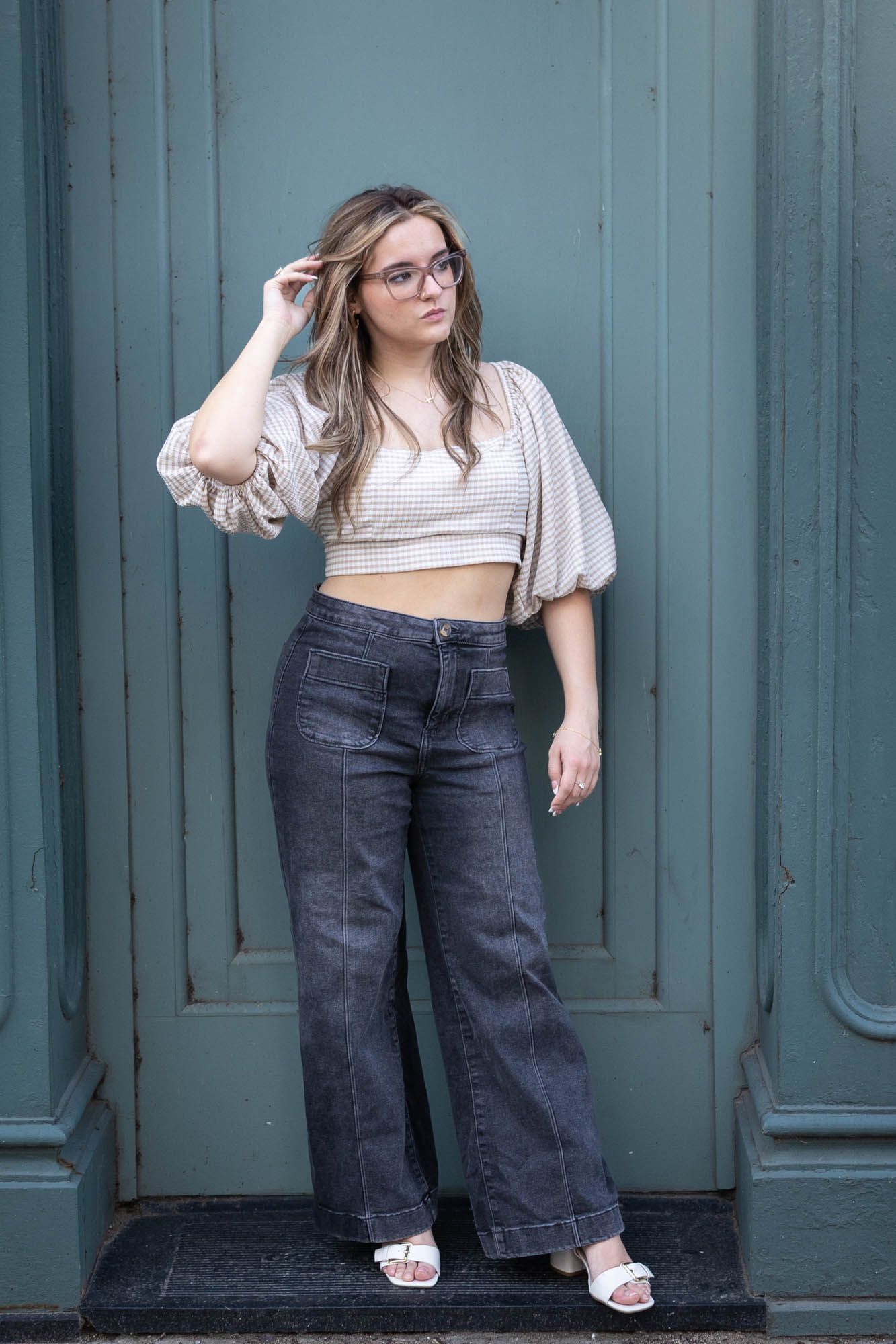 Stylish young woman in glasses, crop top, and wide-leg jeans posing against a teal door.