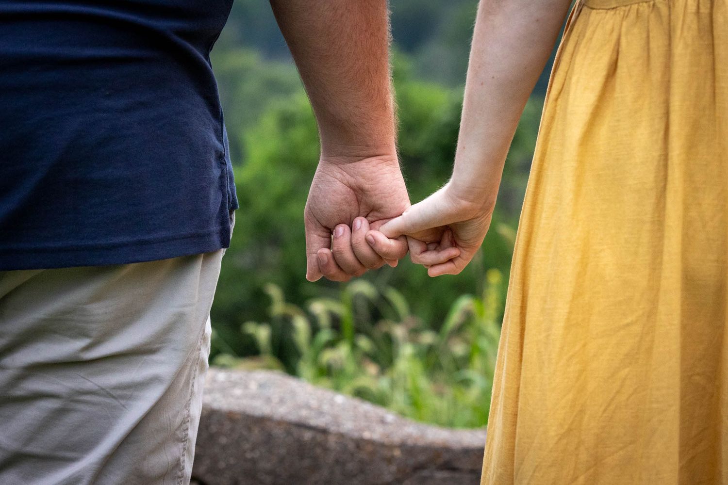 Couple holding hands outdoors, man in blue shirt and woman in yellow dress, with lush green foliage background.
