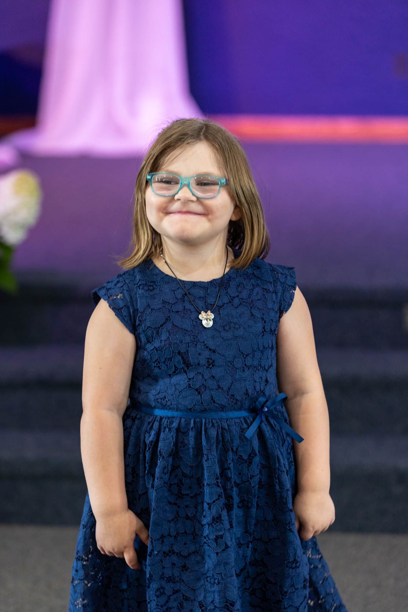 Young girl in navy blue lace dress and teal glasses smiling at a formal event.