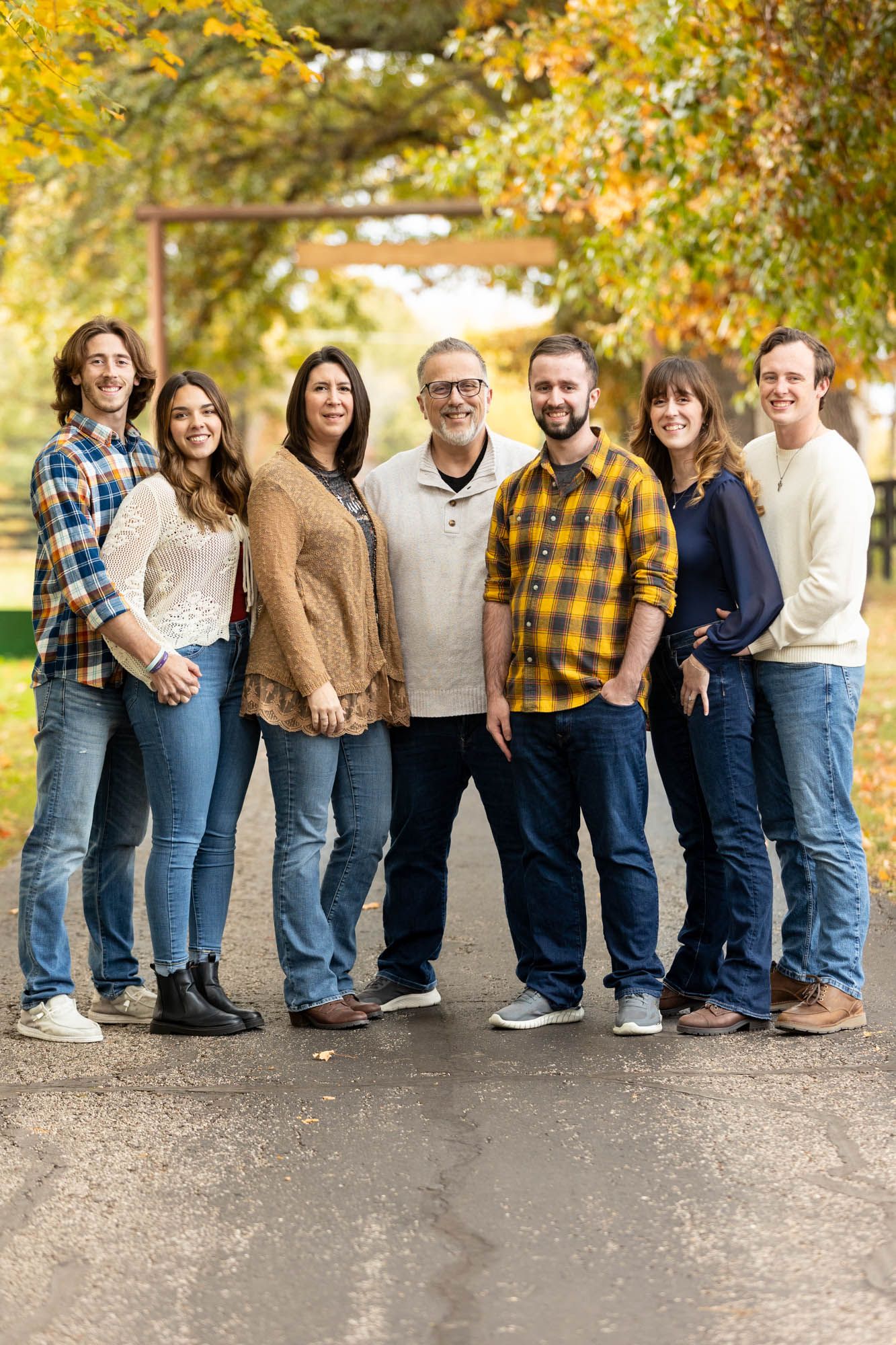 Family of seven posing outdoors in fall, wearing casual flannel and jeans, surrounded by colorful autumn foliage.
