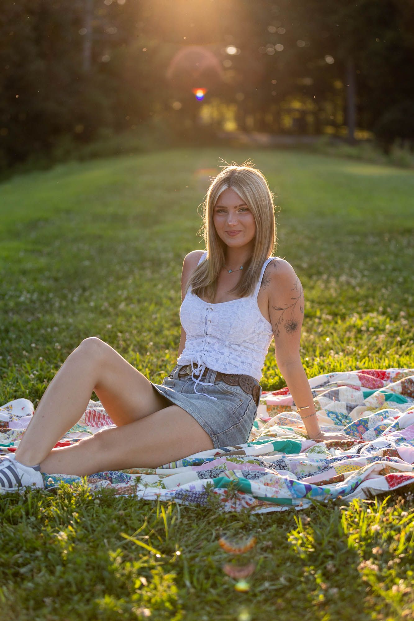 Young blonde woman sitting on a colorful quilt blanket in a sunny park, wearing a white top and denim skirt.