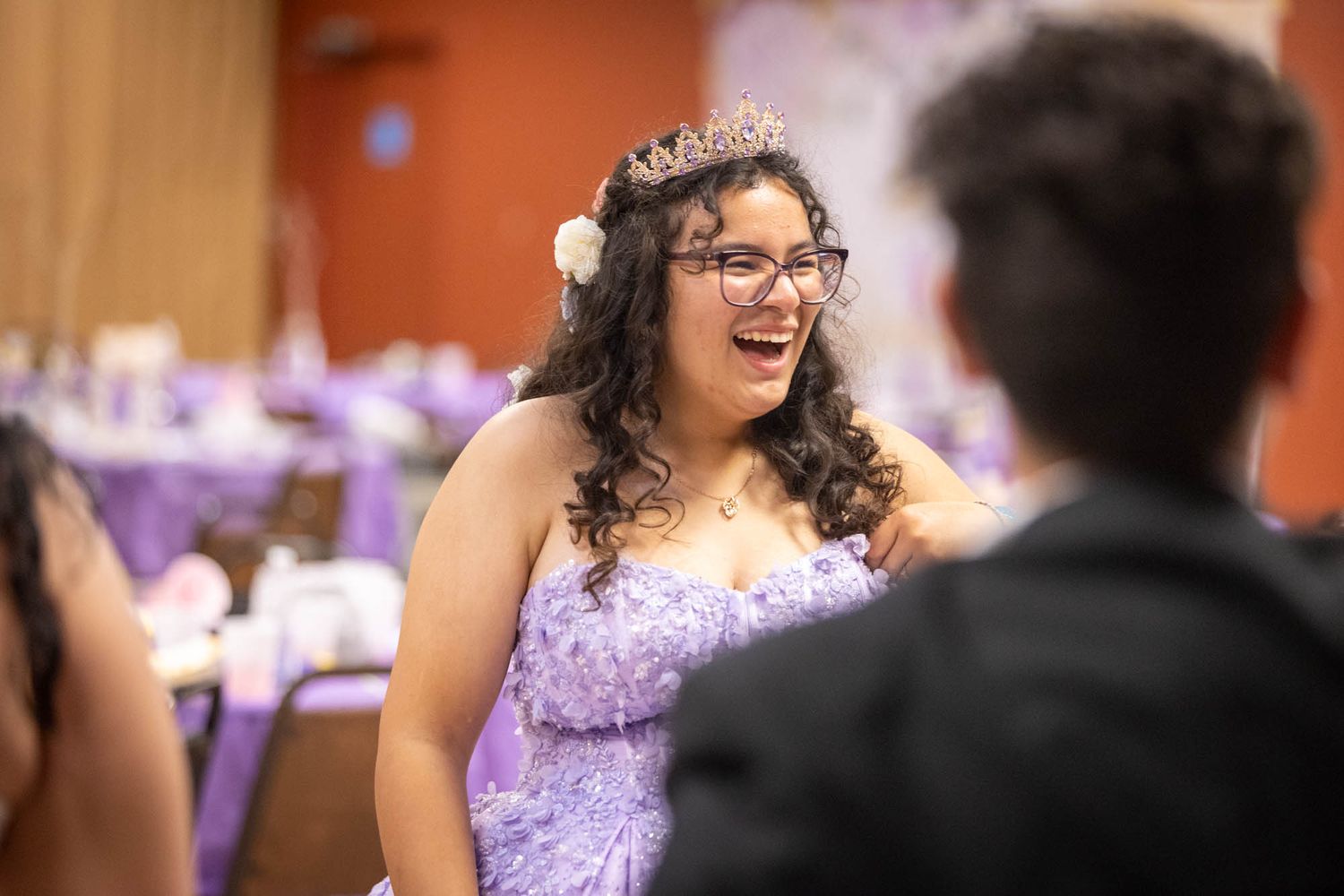 Smiling quinceañera girl wearing tiara and lavender gown laughs with guests at reception.
