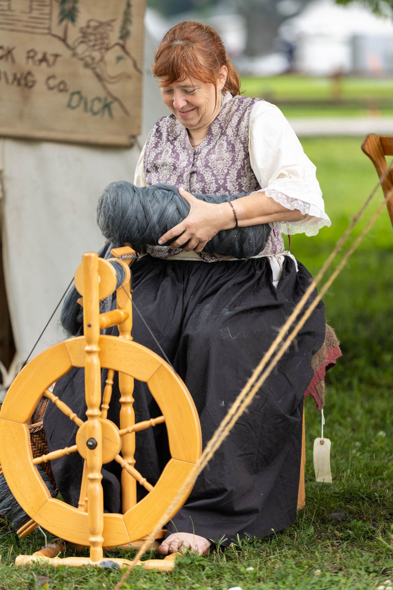A smiling redheaded woman in colonial dress operates a wooden spinning wheel at a historical reenactment.