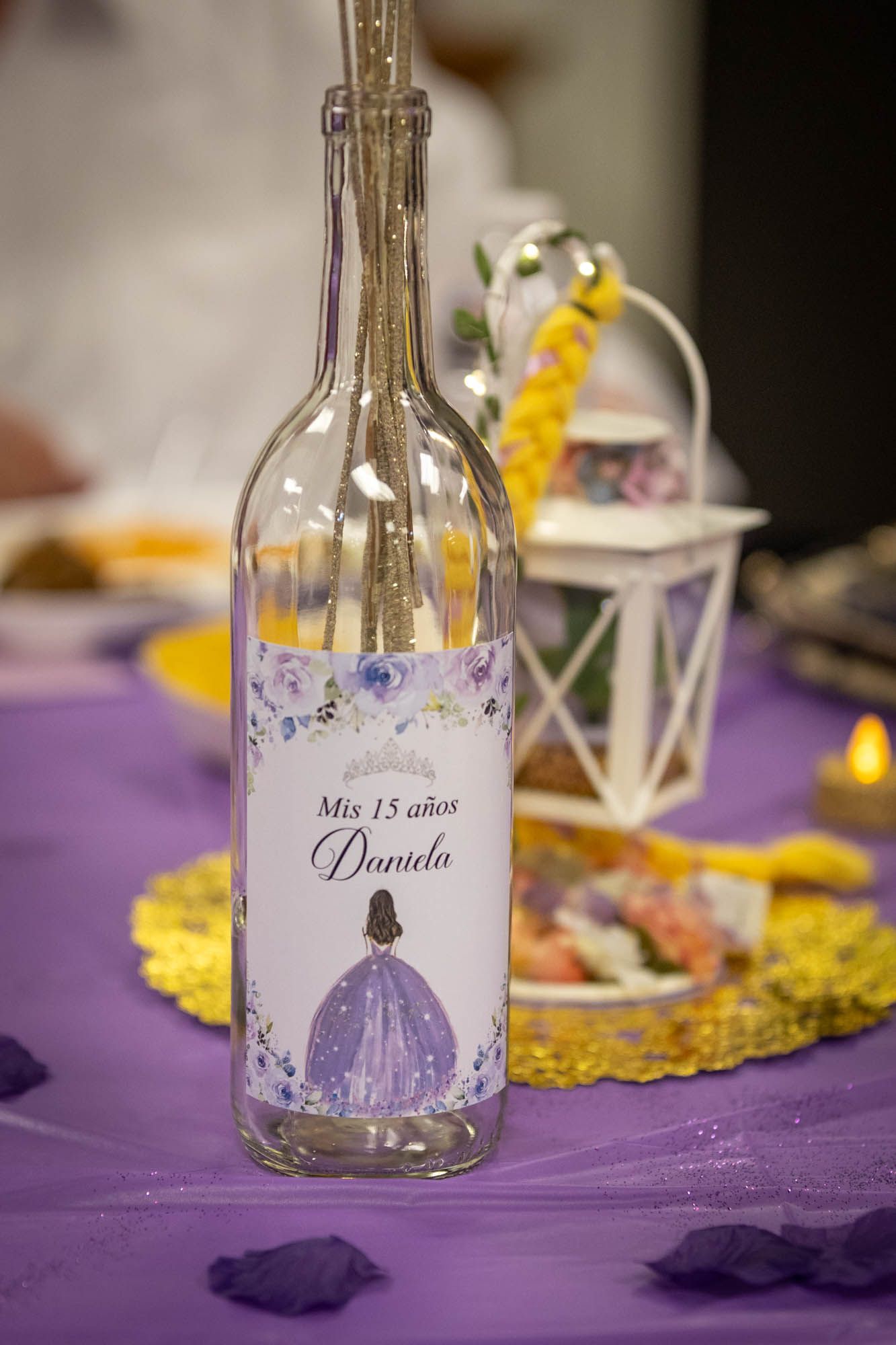 Decorative bottle labeled Mis 15 años Daniela on a purple table at a quinceañera celebration.
