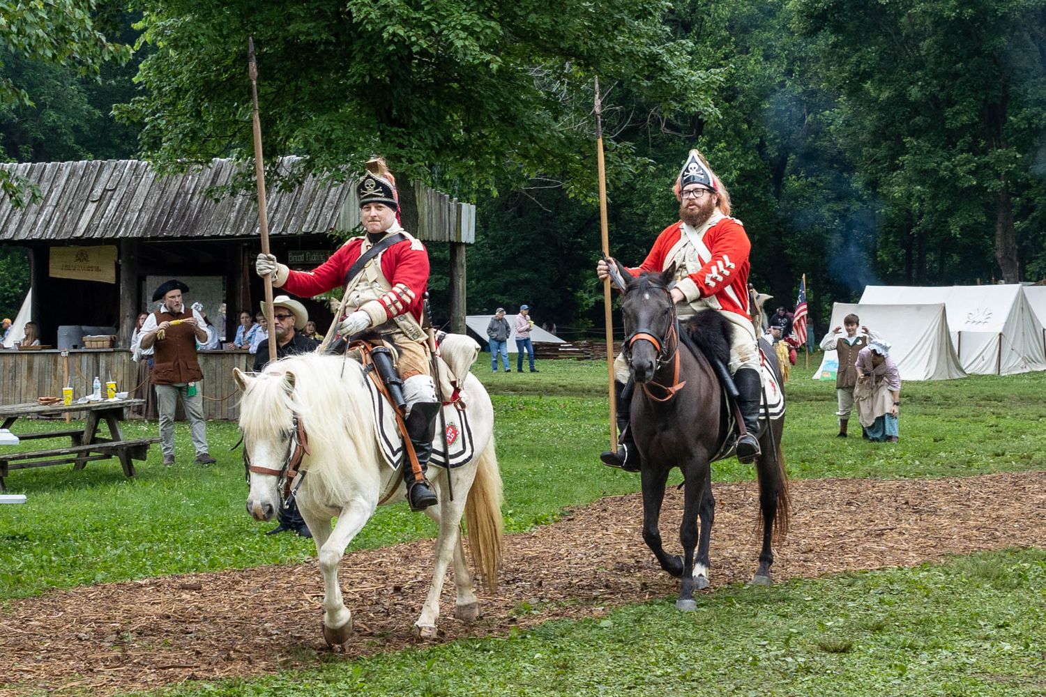 Two riders in red British colonial military uniforms on horseback carrying spears at a historical reenactment.